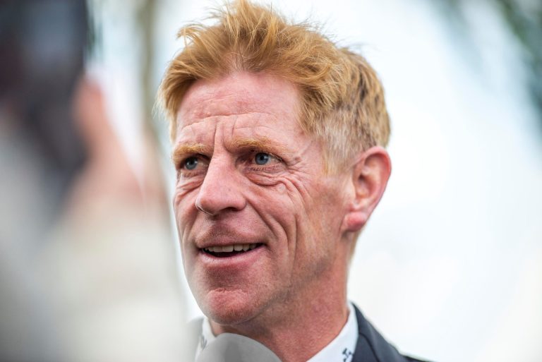 Porträtfoto des Springreiters Marcus Ehning mit rötlich-blonden Haaren in formeller Reitkleidung, der in ein Mikrofon spricht. Der Hintergrund ist unscharf und deutet auf eine Veranstaltung im Freien hin.