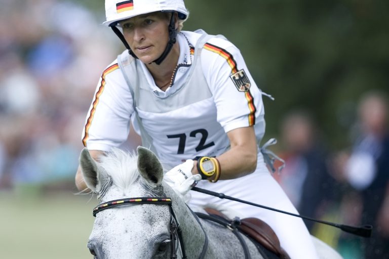 Eventing rider Ingrid Klimke in white competition attire with black, red and gold stripes, start number 72 and the German coat of arms on her sleeve rides a grey horse in a cross-country competition. In the background, other people and green countryside can be seen blurred in the distance.