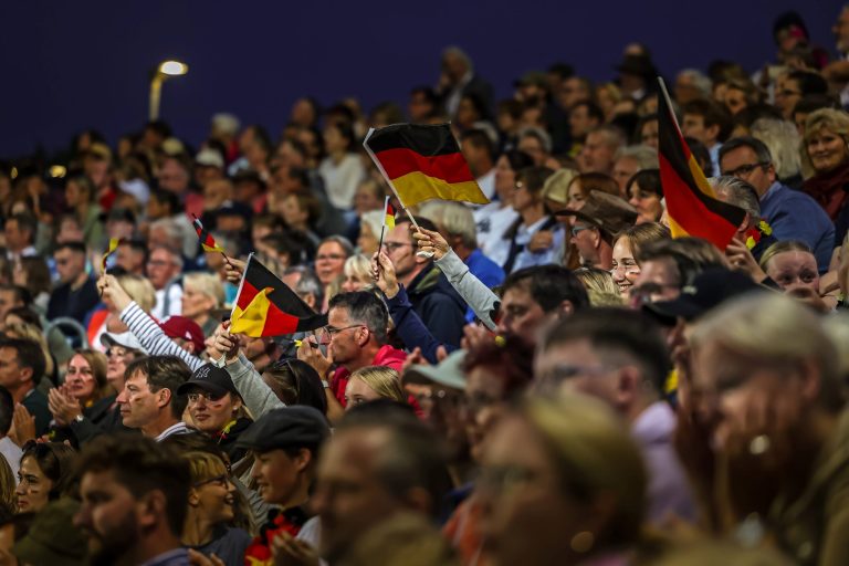 Eine große Menschenmenge sitzt dicht gedrängt auf Tribünen. Viele Zuschauerinnen und Zuschauer schwenken Deutschland-Fähnchen und applaudieren, die Stimmung wirkt ausgelassen und feierlich. Im Hintergrund ist der Himmel bereits dunkel.