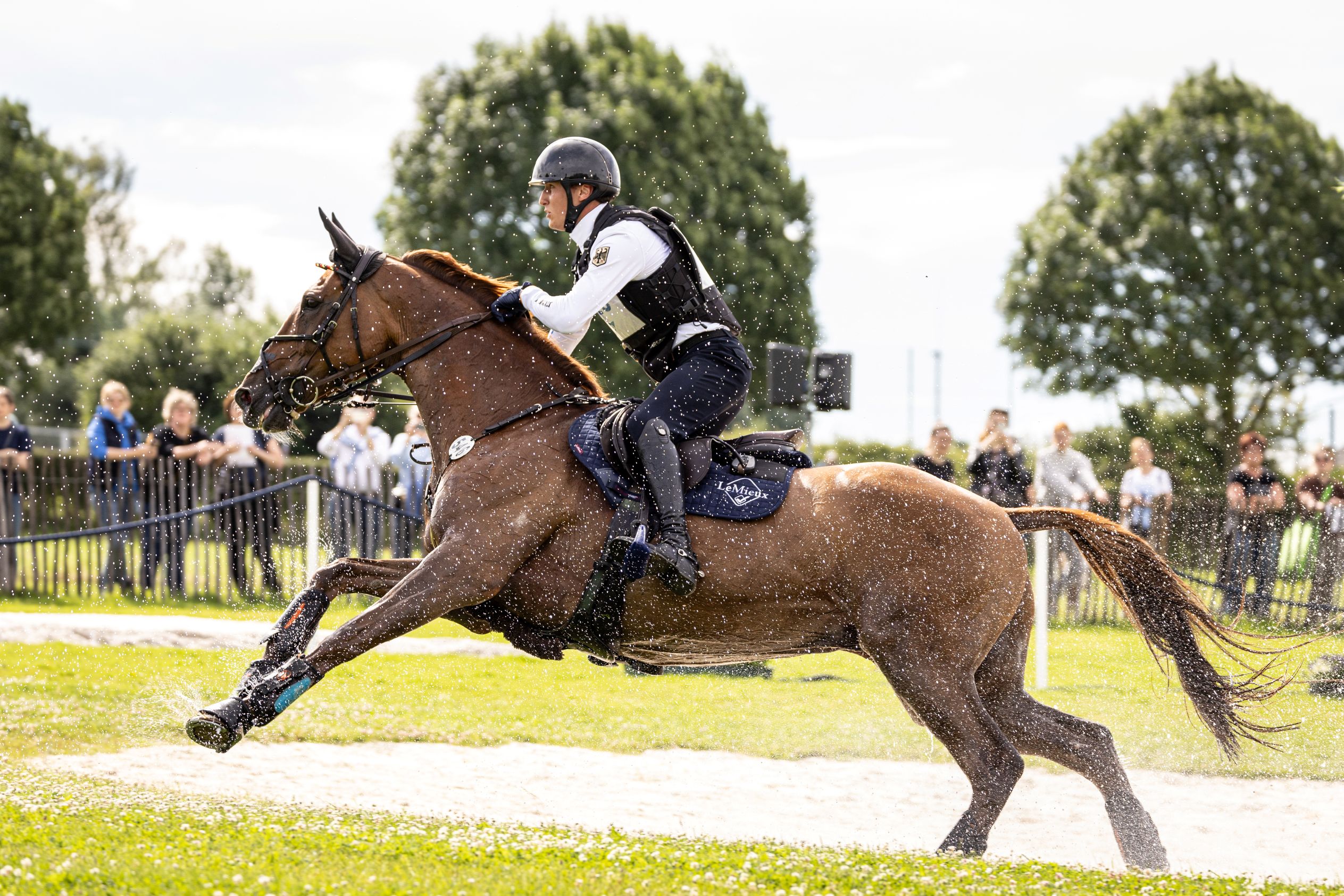 An Eventing rider wearing a protective west and helmet rides a bay horse through a water obstacle, splashing water in all directions. In the background, spectators stand behind a barrier, trees and a bright sky can be seen.