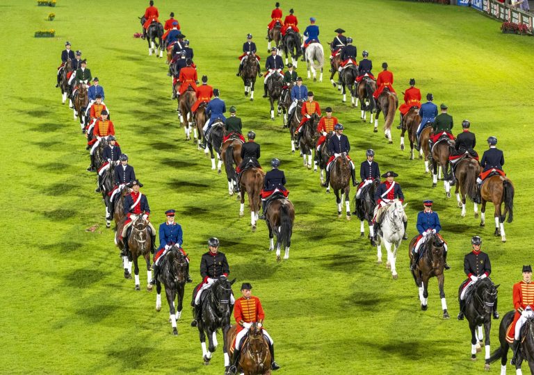 Eine große Gruppe von Reitern in traditioneller Kleidung auf einem grünen Rasenplatz. Die Reiter tragen verschiedene Uniformen in Rot, Blau, Schwarz und Weiß und reiten in mehreren Reihen nebeneinander. Die Pferde sind überwiegend braun oder schwarz, mit einigen weißen Pferden dazwischen. Die Formation erweckt den Eindruck einer feierlichen Parade.