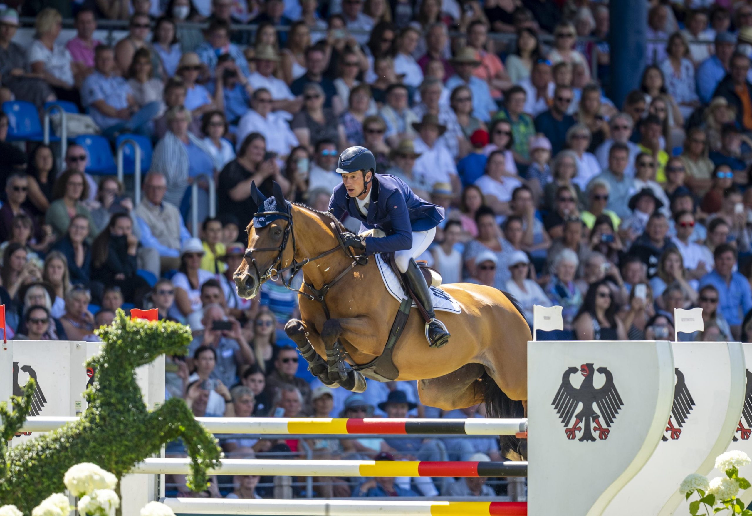 A rider jumps over an obstacle with a horse in a crowded stadium. The obstacle is decorated with the German eagle and in the foreground is a sculpture in the shape of a horse made of plants.