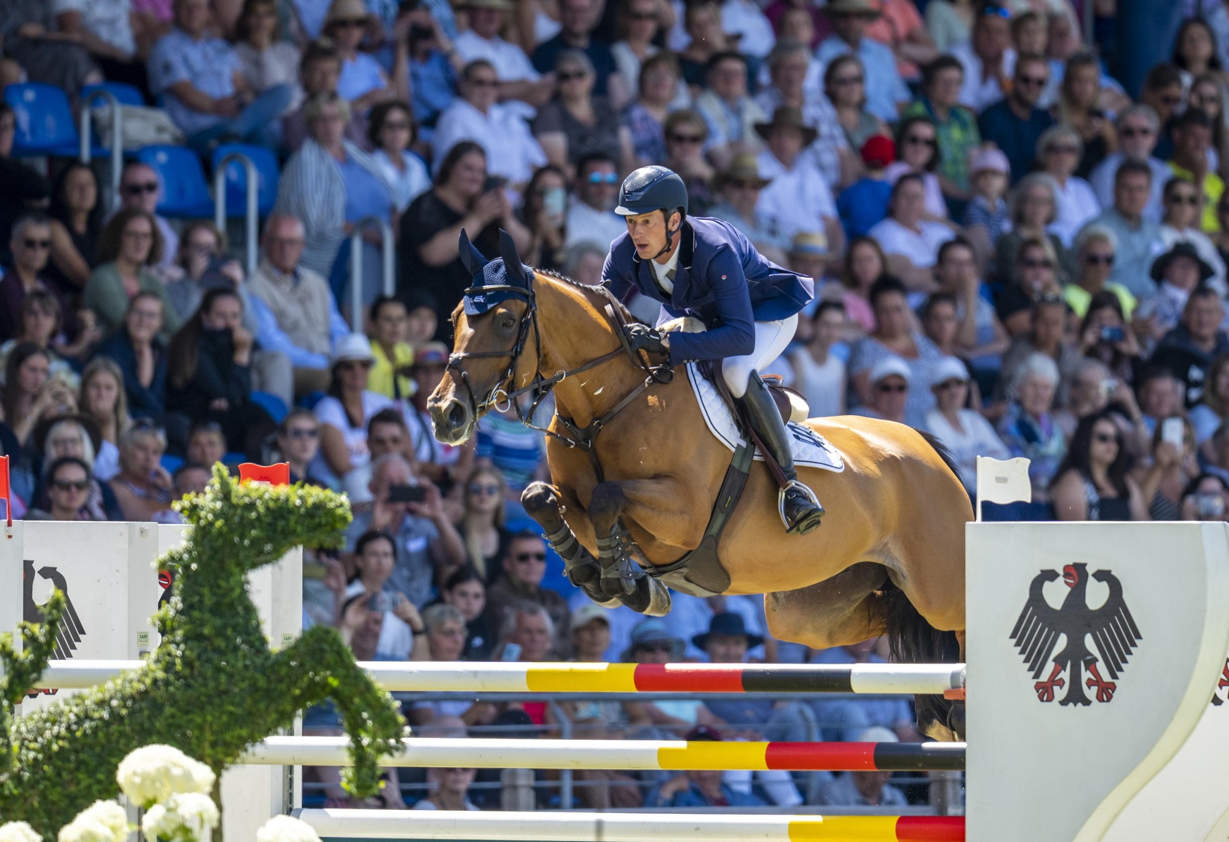 A rider jumps over an obstacle with a horse in a crowded stadium. The obstacle is decorated with the German eagle and in the foreground is a sculpture in the shape of a horse made of plants.