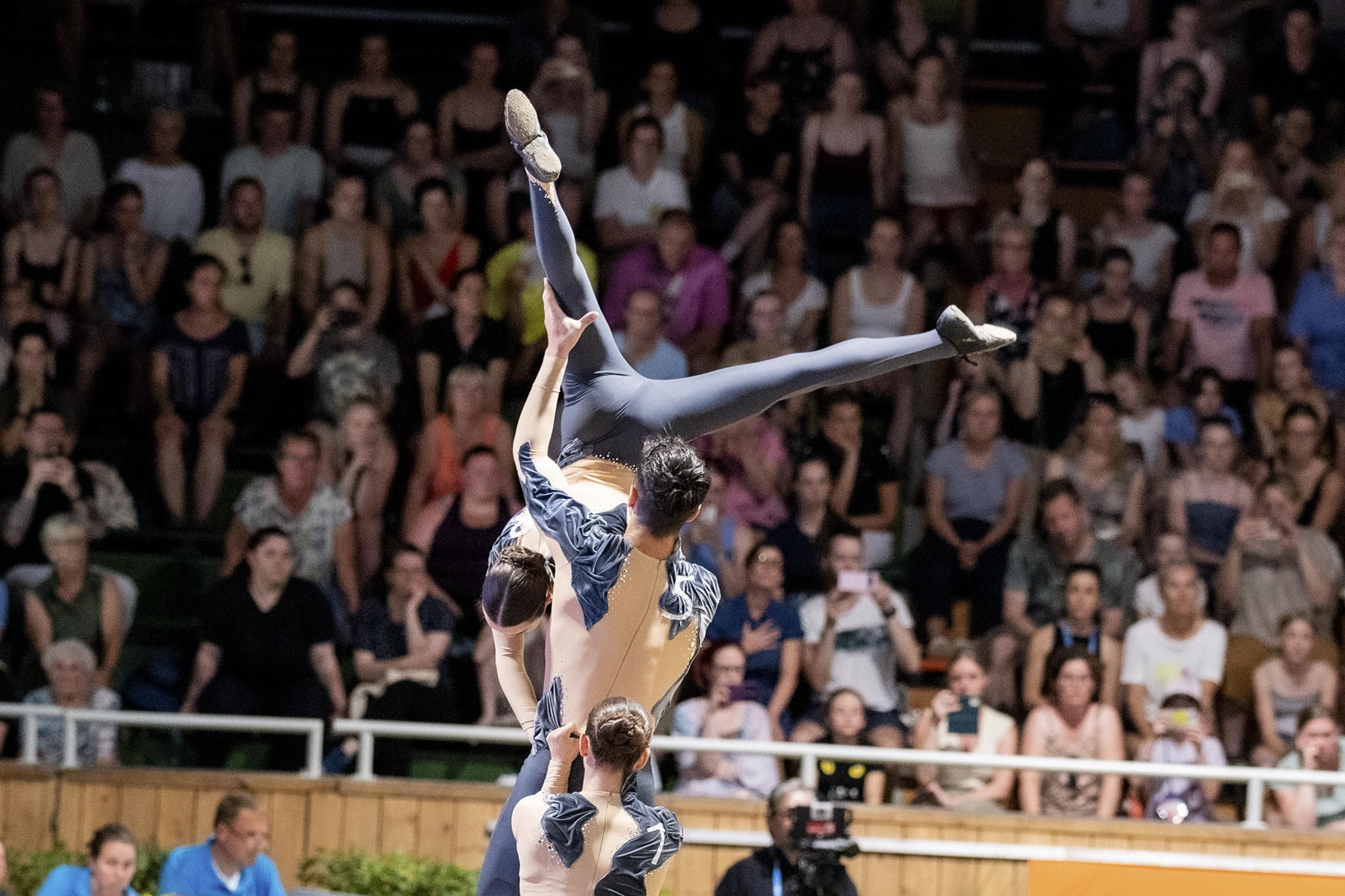 Three vaulters in grey leotards perform an acrobatic lifting figure in an arena. One person lifts a third high above their heads, who hovers horizontally in the air with her legs spread. In the background, a large, blurred audience sits in the stands and watches the performance.
