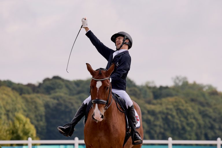Ein Para-Dressurreiter in Reitkleidung und Helm sitzt auf einem braunen Pferd mit weißer Blesse. Der Reiter hebt jubelnd eine Gerte in die Luft. Im Hintergrund sind Bäume und ein heller Himmel zu sehen.