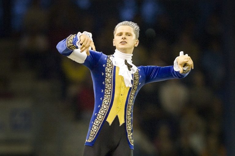 A vaulter stands in a blue and yellow baroque costume on the back of a horse. The costume is inspired by the composer Mozart. The scene takes place in an illuminated arena in front of a blurred audience.