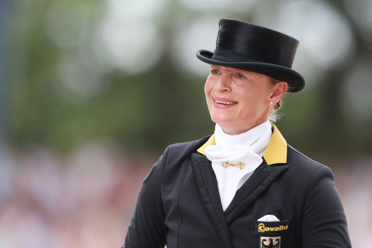 Isabell Werth in classic dressage attire. She is wearing a black tailcoat with gold accents, a white plastron tie and a top hat. The federal eagle can be seen on her breast pocket. The background is blurred, suggesting an outdoor event.