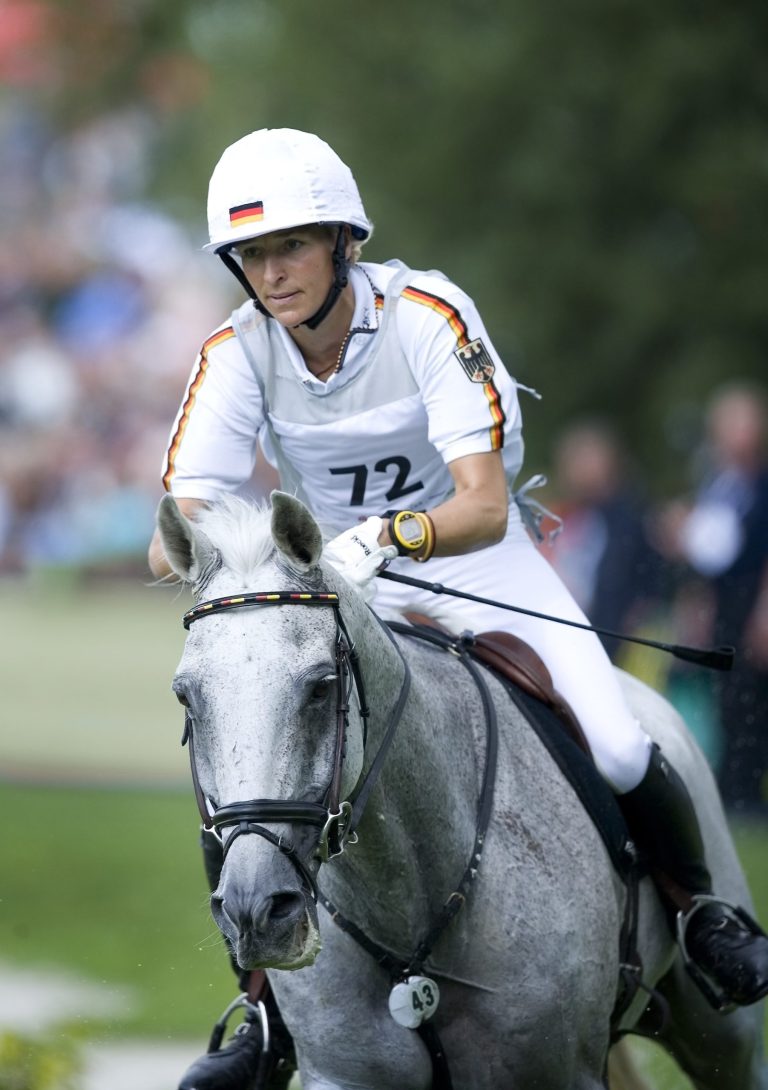 Eventing rider Ingrid Klimke in white competition attire with black, red and gold stripes, start number 72 and the German coat of arms on her sleeve rides a grey horse in a cross-country competition. In the background, other people and green countryside can be seen blurred in the distance.