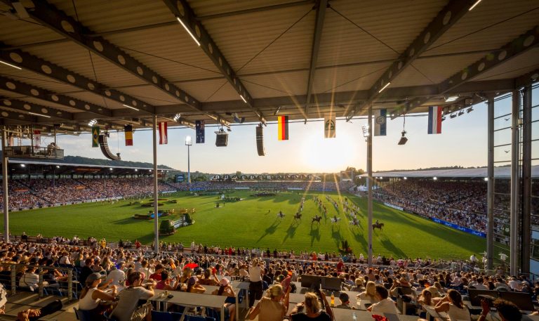 Wide-angle shot of the packed main stadium in Aachen during an opening ceremony in sunny weather. In the foreground, you can see the covered grandstand with spectators, while in the background, numerous horses ride into the stadium. Flags of various nations hang from the grandstand roofs.