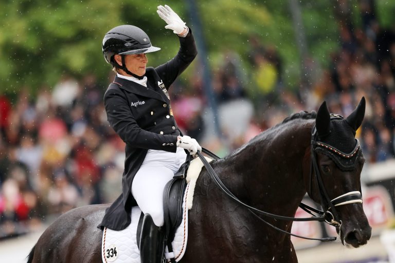 A dressage rider in black competition attire and a helmet is riding a black horse with a white pad and raises her right hand in greeting. In the background, spectators can be seen in the stands and it is drizzling.