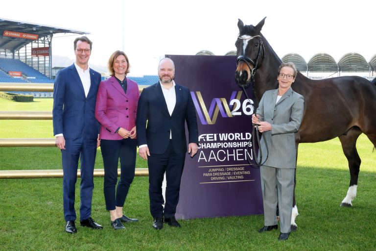Four people and a horse stand next to a large sign reading “FEI World Championships Aachen 2026” on the grass in the stadium. The people are dressed formally, and one person is holding the horse by the halter. Stadium grandstands and seating are visible in the background.