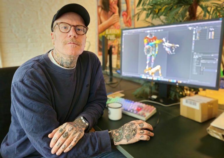 A tattooed man wearing glasses and a cap sits at a desk and looks into the camera. In front of him is a large monitor displaying a colorful 3D graphic. On the desk are a keyboard, a mug, and a small package.