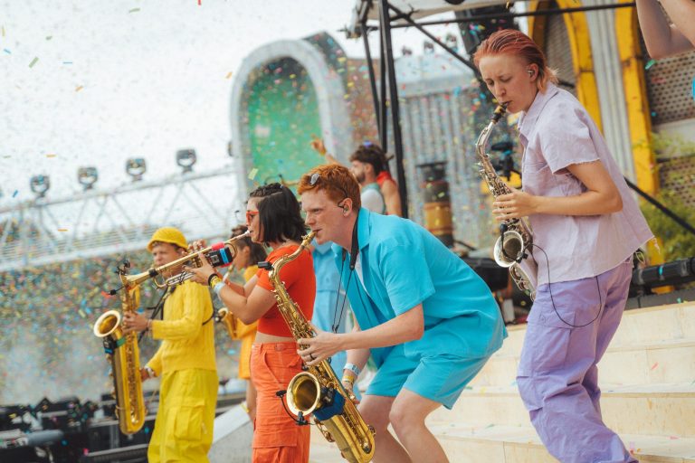 A colourfully dressed brass band performing energetically on a festival stage. Several people are playing saxophones and trumpets, whilst confetti is flying through the air in the background. The musicians are wearing yellow, orange, blue and purple outfits. The atmosphere is dynamic and festive.