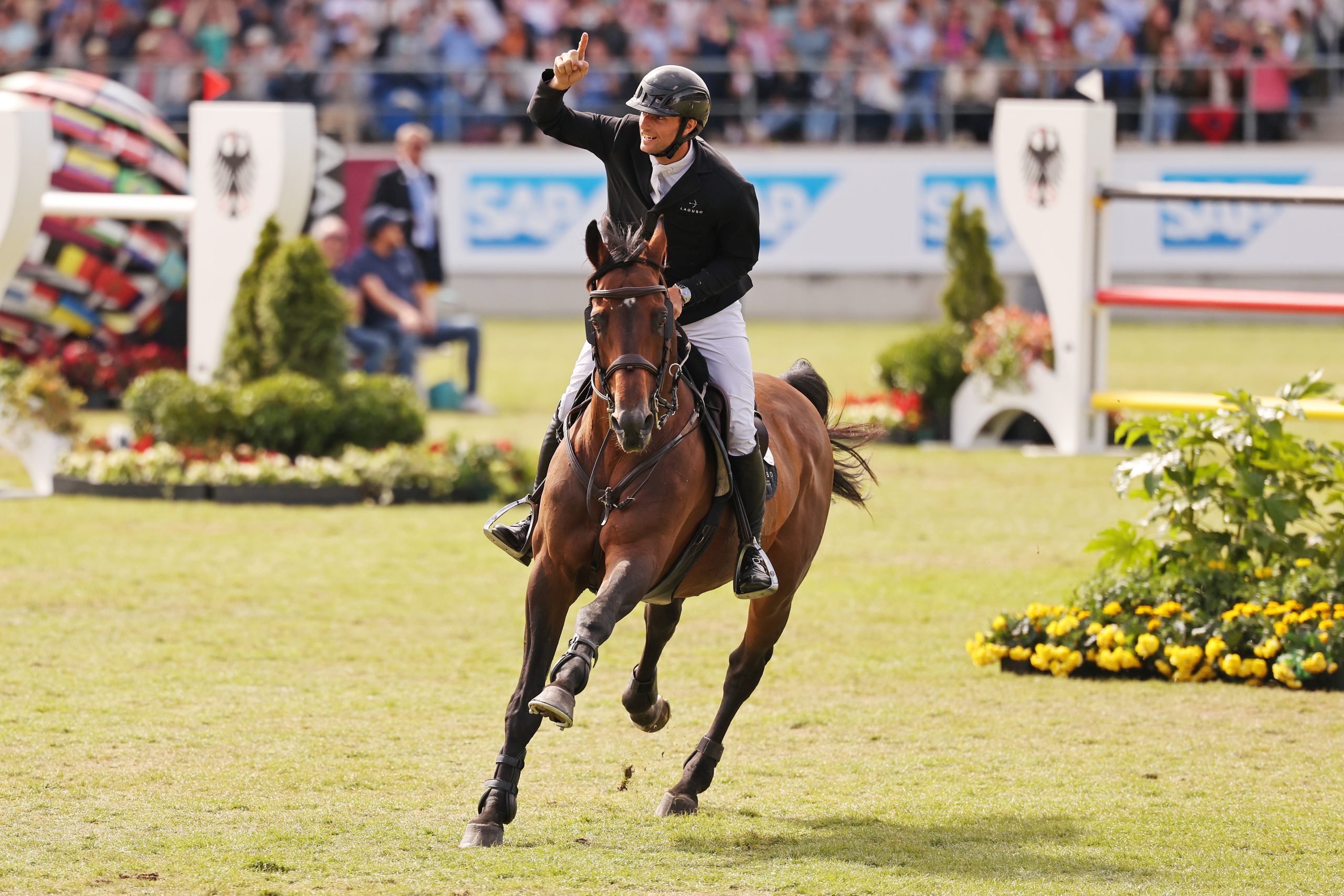 Show jumper on a brown horse gallops victoriously across a grass arena, raising his right arm with one finger pointed upward; in the background are fences with a federal eagle emblem, sponsor boards, and packed spectator stands.