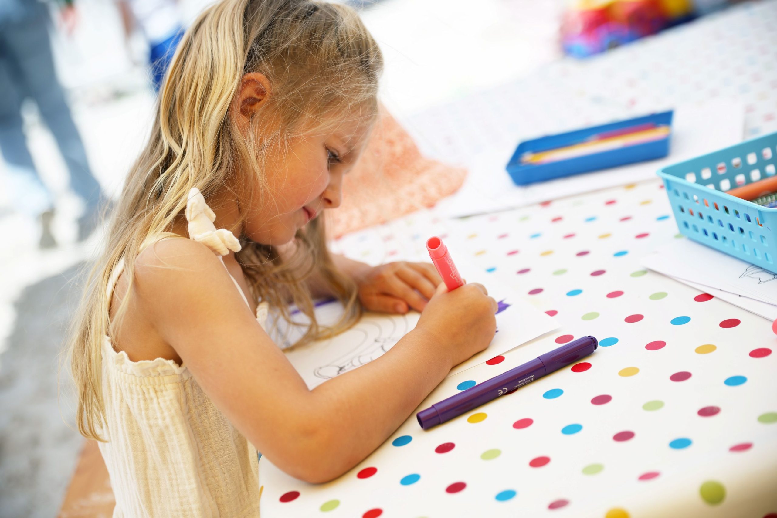 A little blonde girl is sitting at a table covered with a colourful polka-dot tablecloth, intently drawing with a pink felt-tip pen; next to her are more pens and craft supplies.