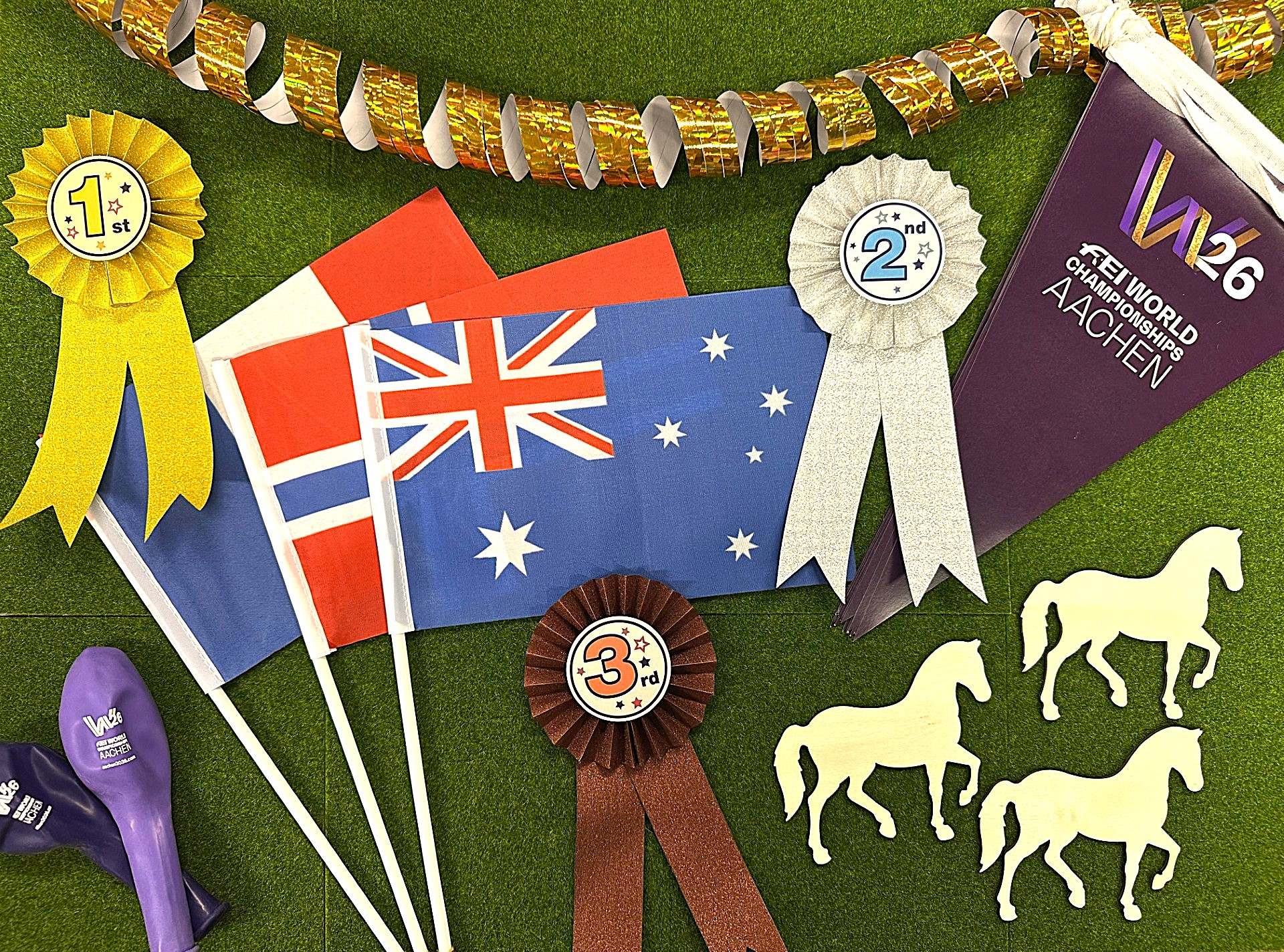 Colourful decorations on a green background for the equestrian event in Aachen: in the centre are small flags of Norway, Switzerland and Australia. A gold garland hangs at the top. On the left is a yellow winner’s ribbon bearing the inscription ‘1st’, at the bottom a brown ribbon with ‘3rd’, and on the right a large white ribbon with ‘2nd’. In the top right-hand corner is a purple pennant bearing the inscription ‘W26 FEI World Championships Aachen’. At the bottom left are two purple balloons with the same logo, and at the bottom right four light-coloured horse silhouettes.