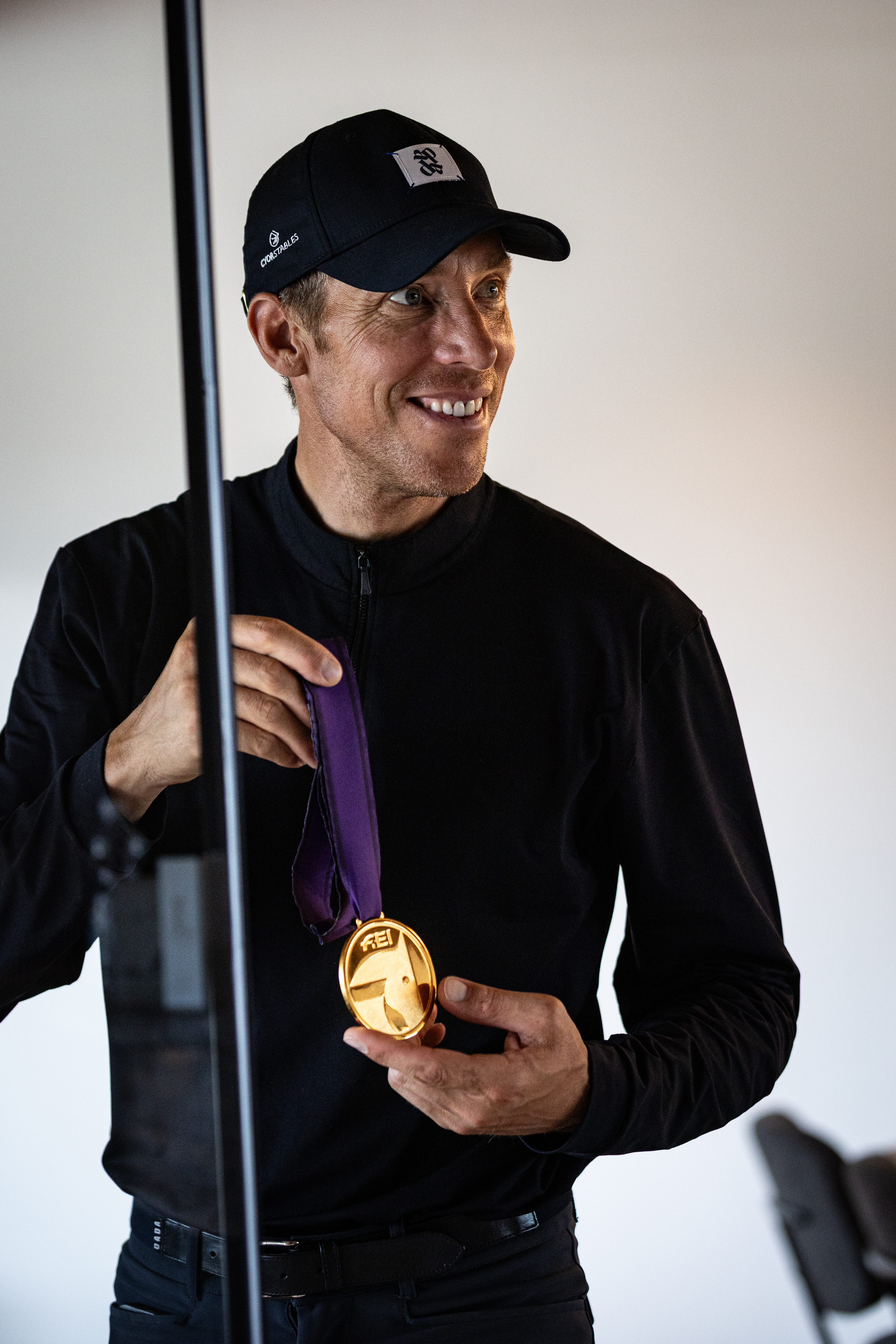 Lächelnder Mann in schwarzer Kappe und dunkler Kleidung hält eine goldene FEI-Medaille an einem violetten Band vor hellem, neutralem Hintergrund in der Hand.