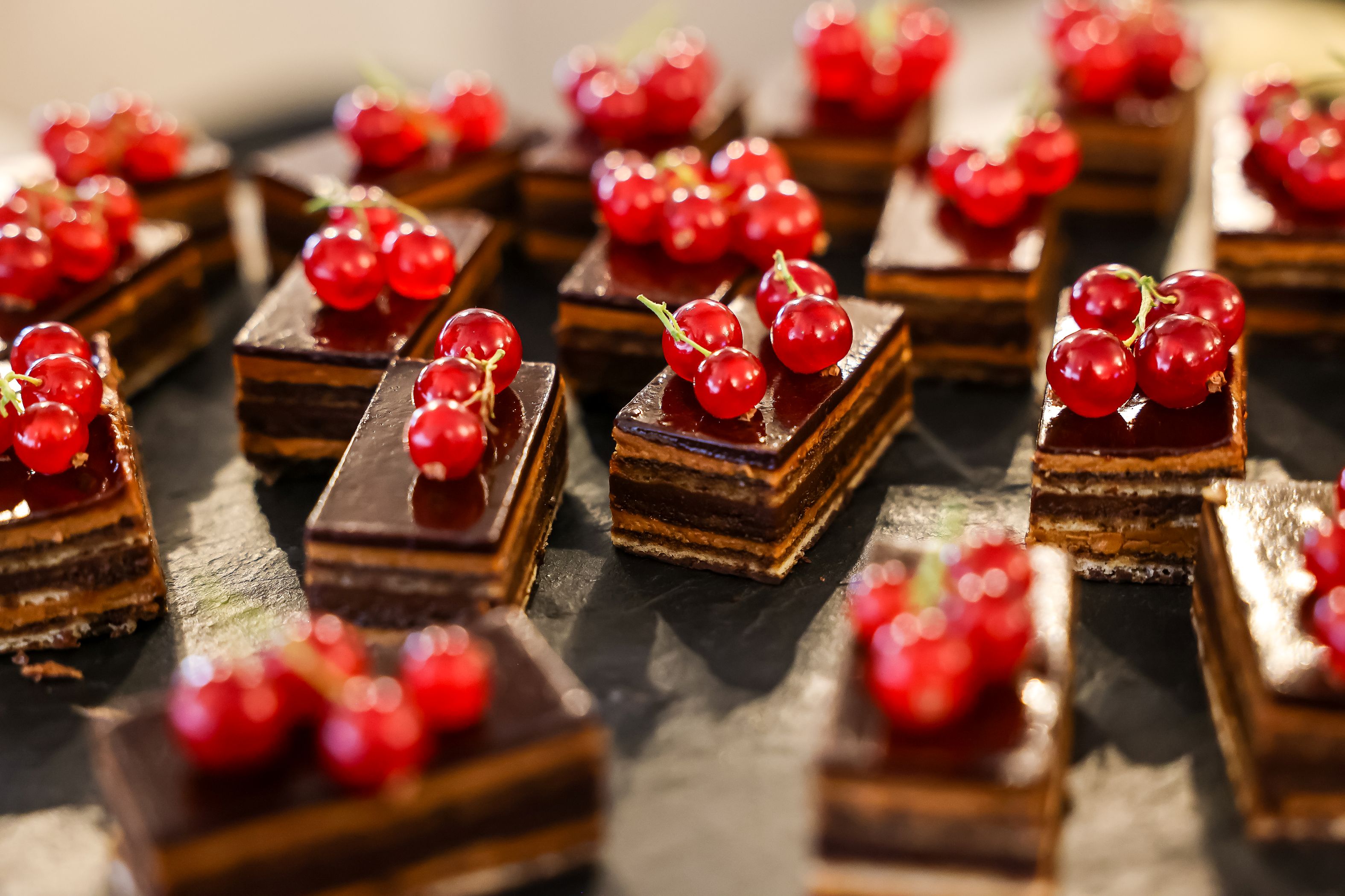 Nahaufnahme von kleinen, rechteckigen Schoko-Schnitten mit glänzender Glasur, die jeweils mit roten Johannisbeeren garniert sind.