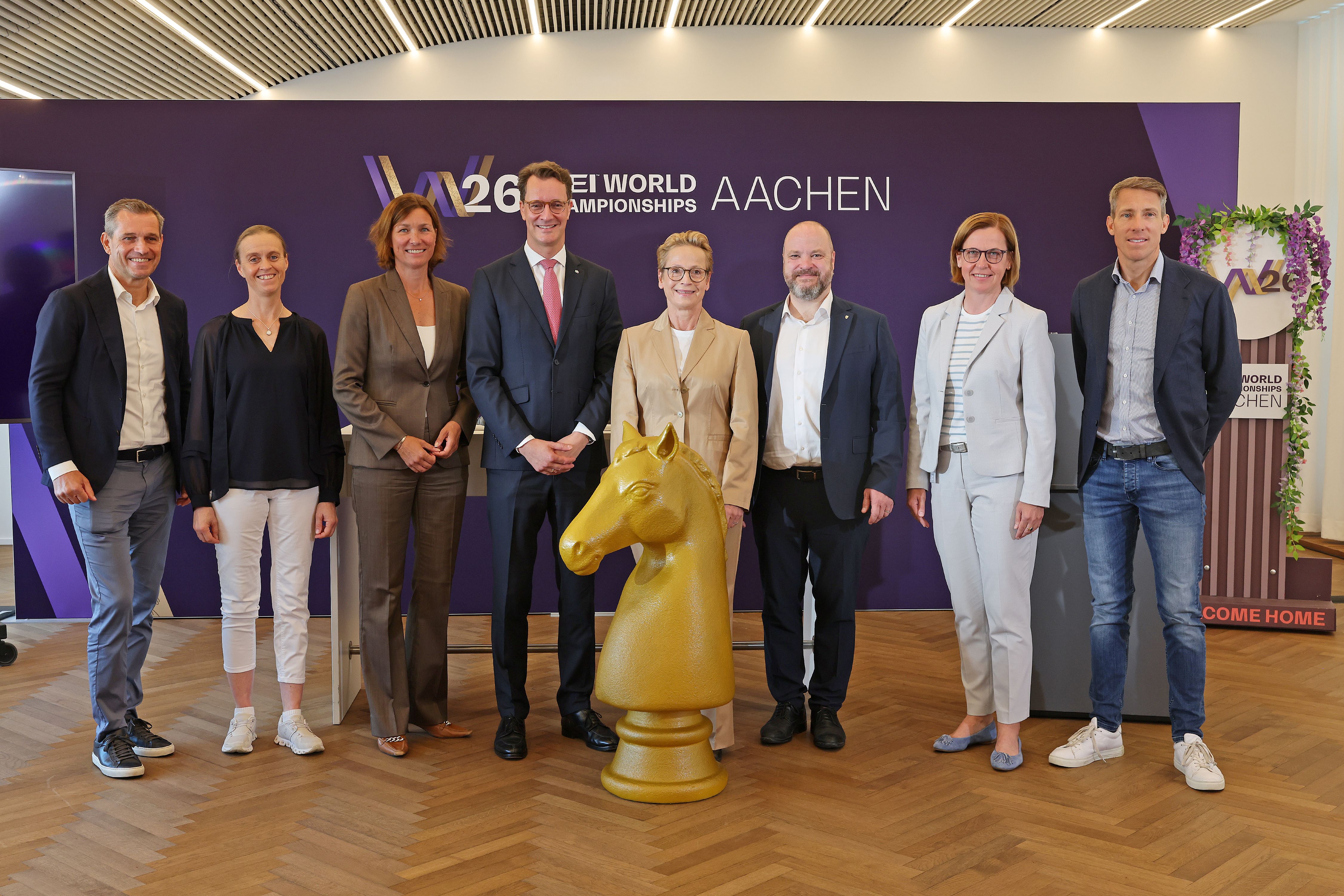 Acht Personen stehen in einer Reihe vor einer lila Wand mit dem Schriftzug ‚W26 FEI World Championships Aachen‘; in der Mitte vor der Gruppe steht eine große goldene Pferdekopf-Skulptur auf einem Holzfußboden.