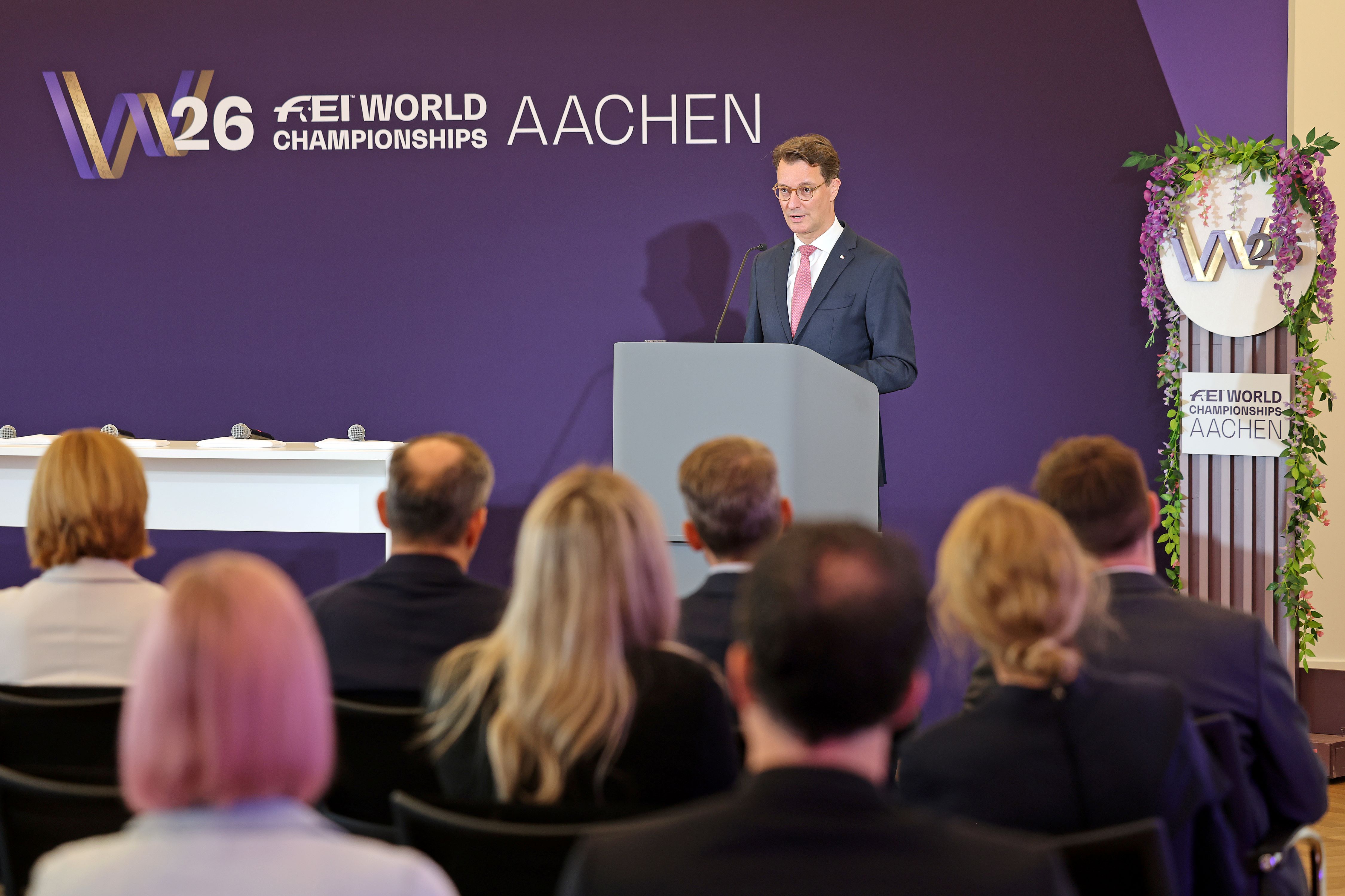 Ein Redner steht an einem grauen Pult vor einer lila Bühnenwand mit dem Schriftzug ‚W26 FEI World Championships Aachen‘ und spricht in ein Mikrofon, im Vordergrund sitzt das Publikum in mehreren Reihen.
