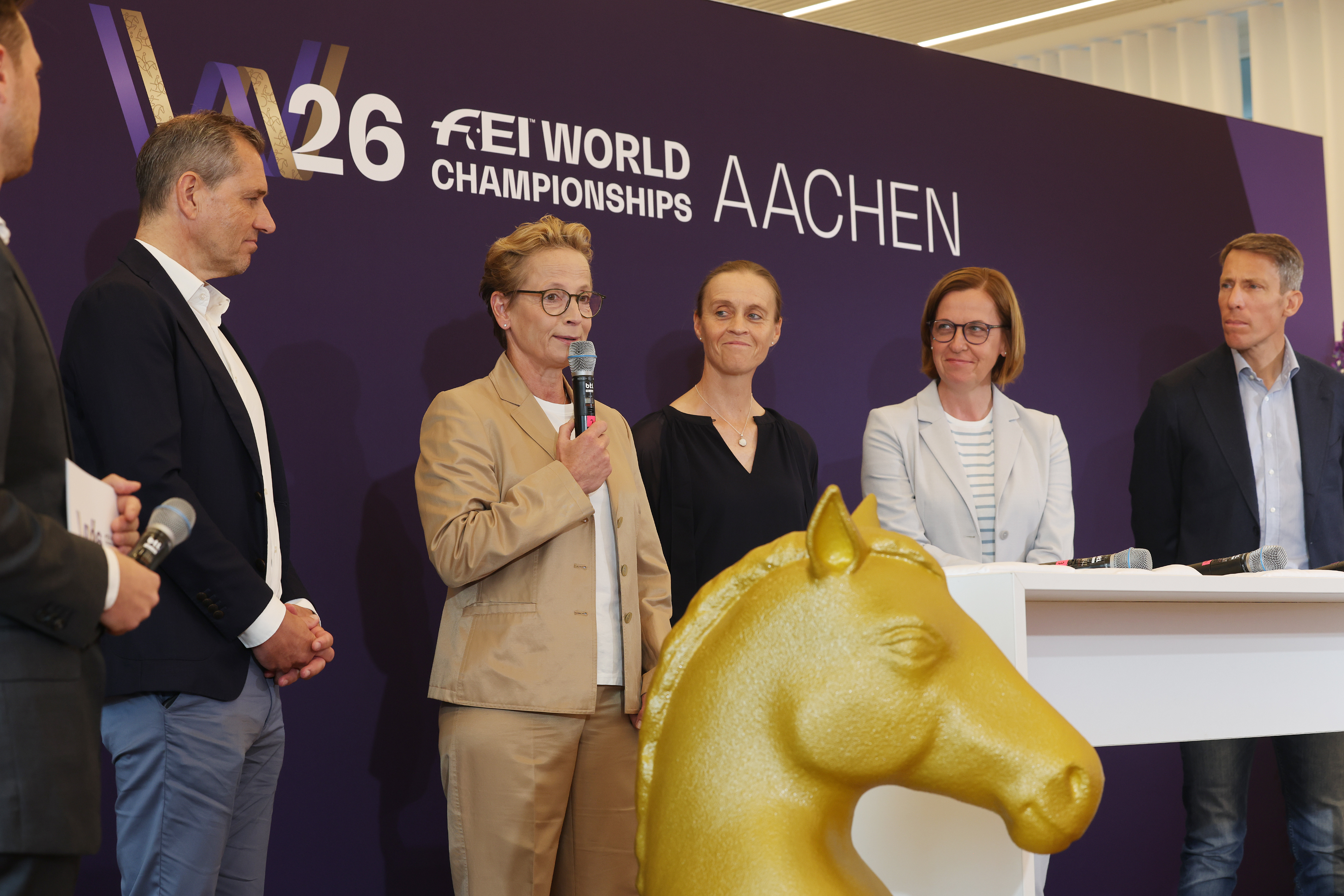 Mehrere Personen stehen vor einer lila Wand mit dem Schriftzug ‚W26 FEI World Championships Aachen‘; eine Frau im beigefarbenen Anzug spricht mit einem Mikrofon, daneben stehen weitere Gesprächsteilnehmende, im Vordergrund ist eine große goldene Pferdekopf-Skulptur zu sehen.