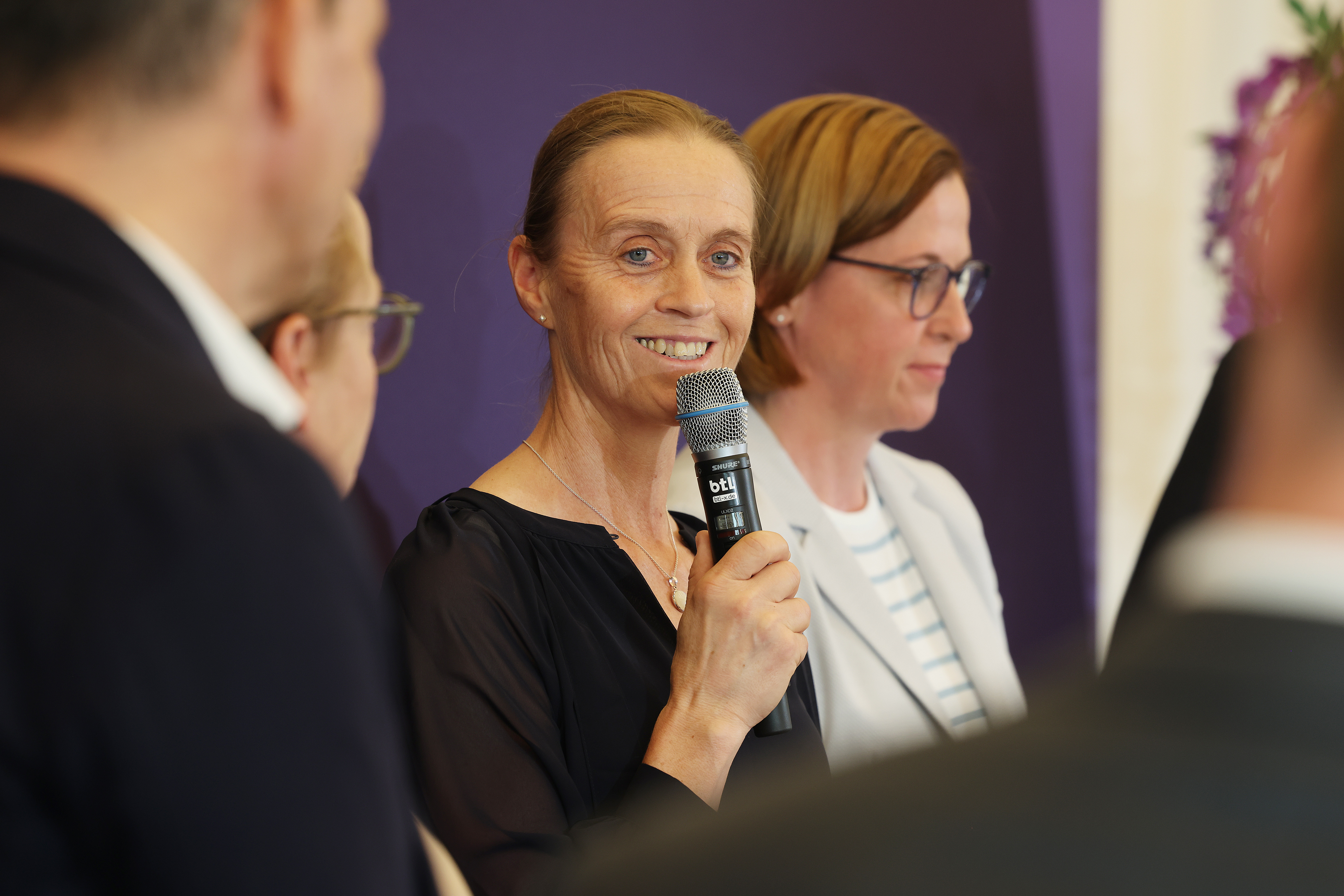 Eine lächelnde Frau hält ein Mikrofon und spricht, im Hintergrund stehen weitere Personen vor einer lila Wand, teilweise unscharf im Vorder- und Hintergrund.