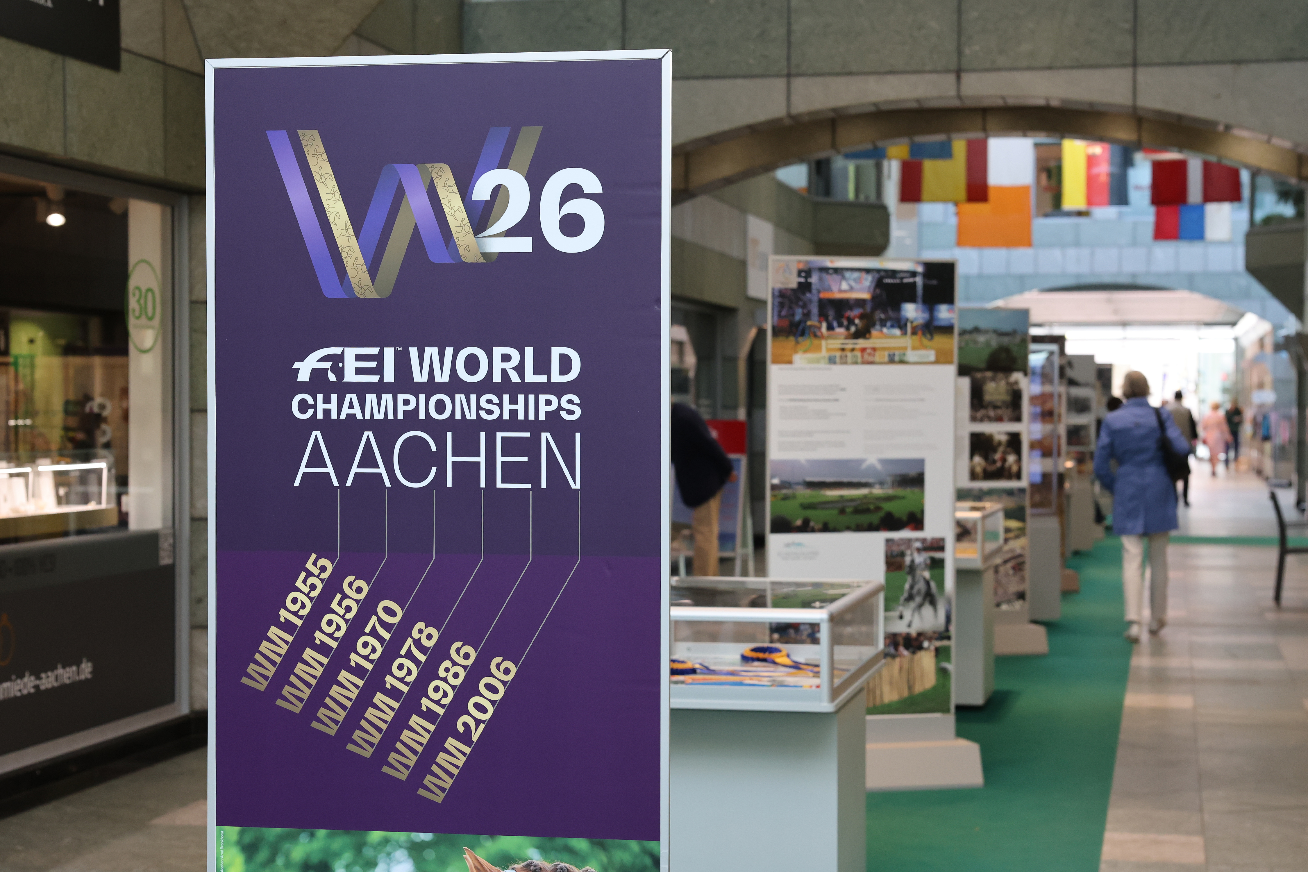 Großes violett-goldenes Roll-up mit dem Logo „W26“ und der Aufschrift „FEI World Championships Aachen“ in einer überdachten Passage; darunter sind frühere WM-Jahre aufgelistet. Im Hintergrund verläuft eine Ausstellung mit Informationstafeln und Vitrinen zu den Aachener Reitsport-Weltmeisterschaften, zwischen denen Besucherinnen und Besucher entlanggehen.