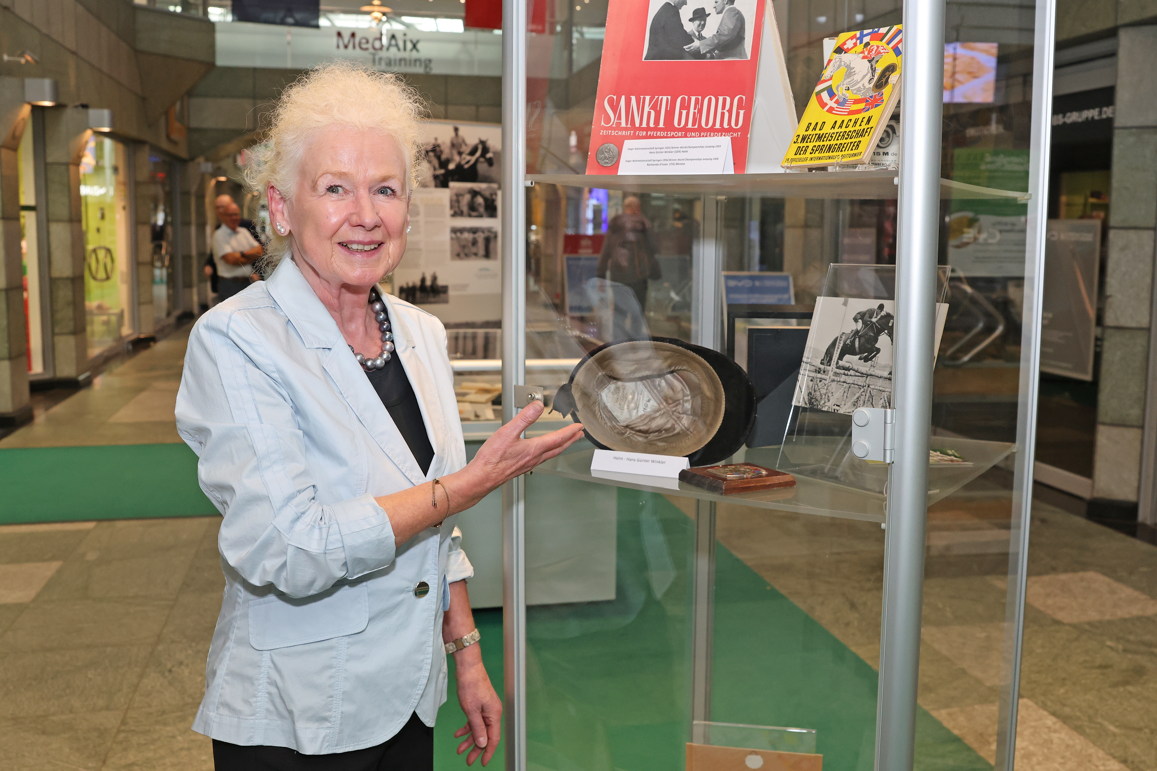 Frau in hellblauem Blazer steht in einer Passage vor einer Glasvitrine einer Reitsport-Ausstellung und lächelt in die Kamera; mit der rechten Hand deutet sie auf ein ausgestelltes einen alten Reithelm, daneben liegen historische Magazine und Schwarz-Weiß-Fotos aus dem Pferdesport.