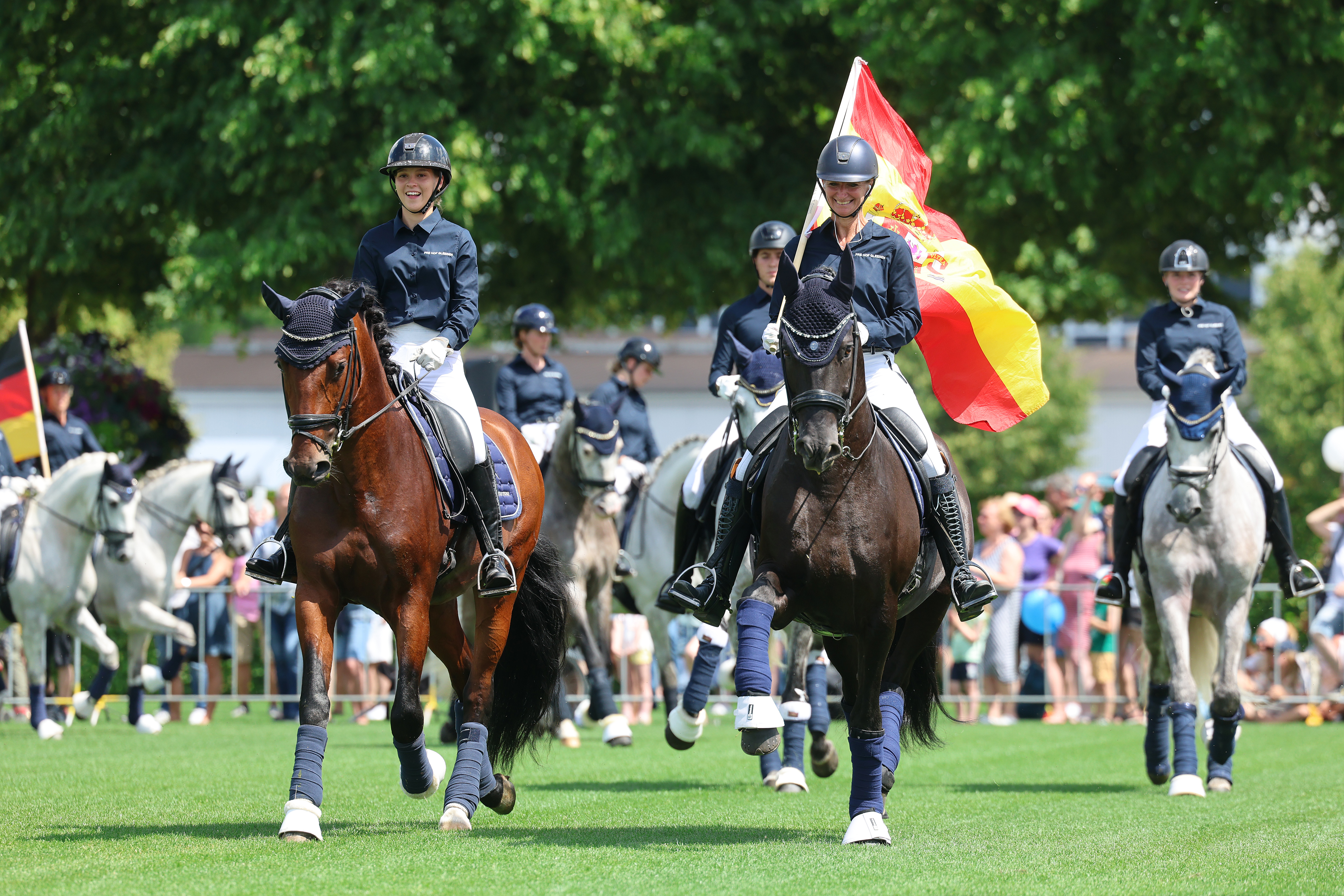 Reiterinnen und Reiter in dunkelblauen Jacken reiten bei Sonnenschein über den grünen Rasen, im Vordergrund trägt eine Reiterin auf einem dunklen Pferd eine große rot-gelb-rote Flagge, im Hintergrund stehen weitere Pferde sowie Zuschauer.