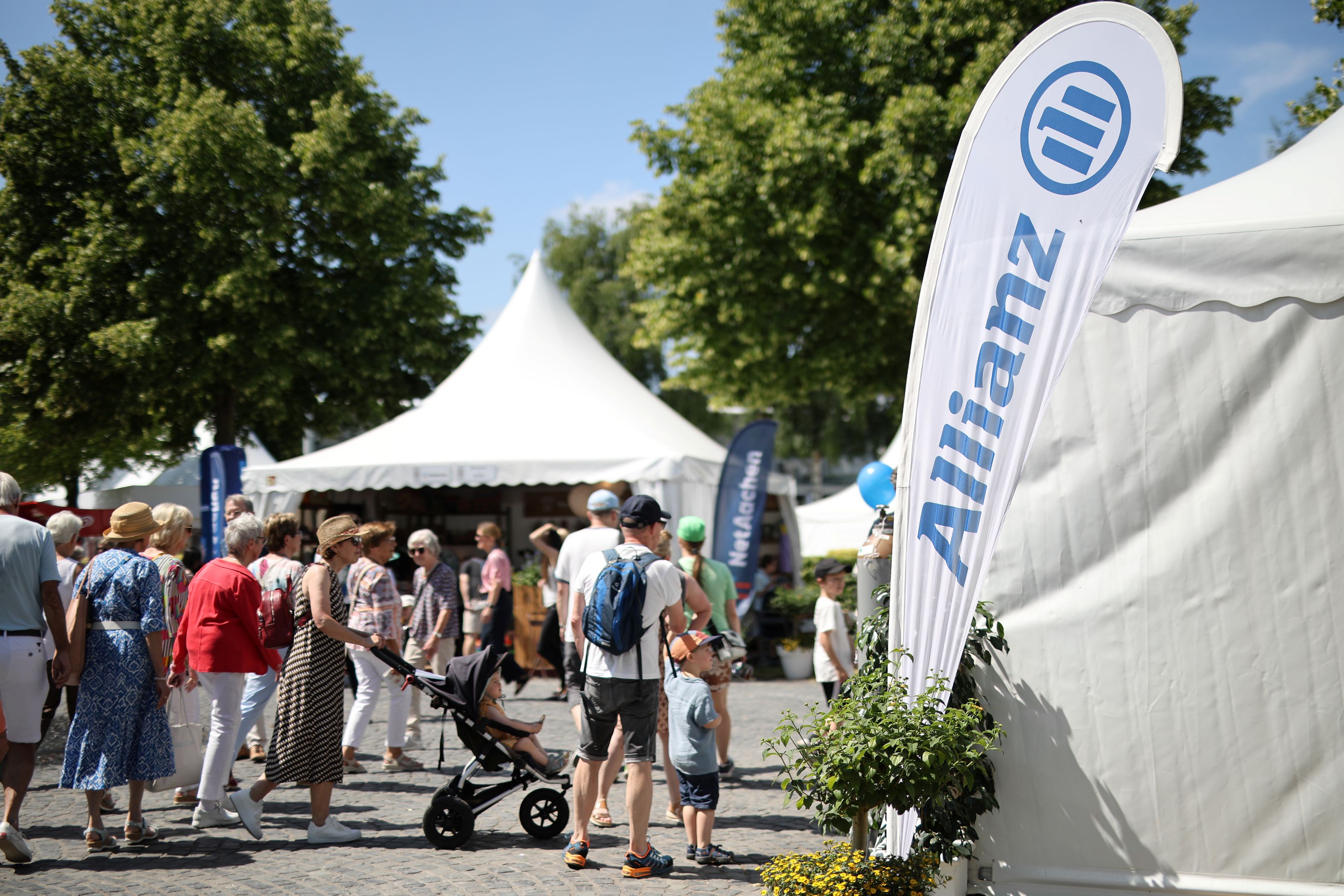 Menschen schlendern bei sonnigem Wetter über ein Außengelände mit weißen Pagodenzelten; im Vordergrund steht ein Kind im Buggy, daneben ein Mann mit Rucksack, rechts ragt eine weiße Beachflag mit blauem Allianz-Logo in die Höhe.