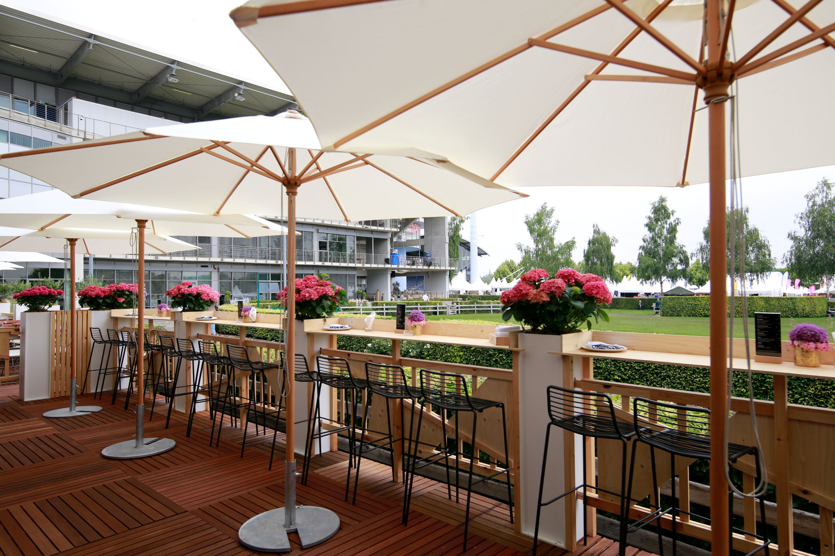 Holzterrasse mit hoher Theke und Barhockern unter großen weißen Sonnenschirmen; auf der Theke stehen Hortensien in Töpfen, im Hintergrund sieht man das Turniergelände und die Geschäftsstelle.