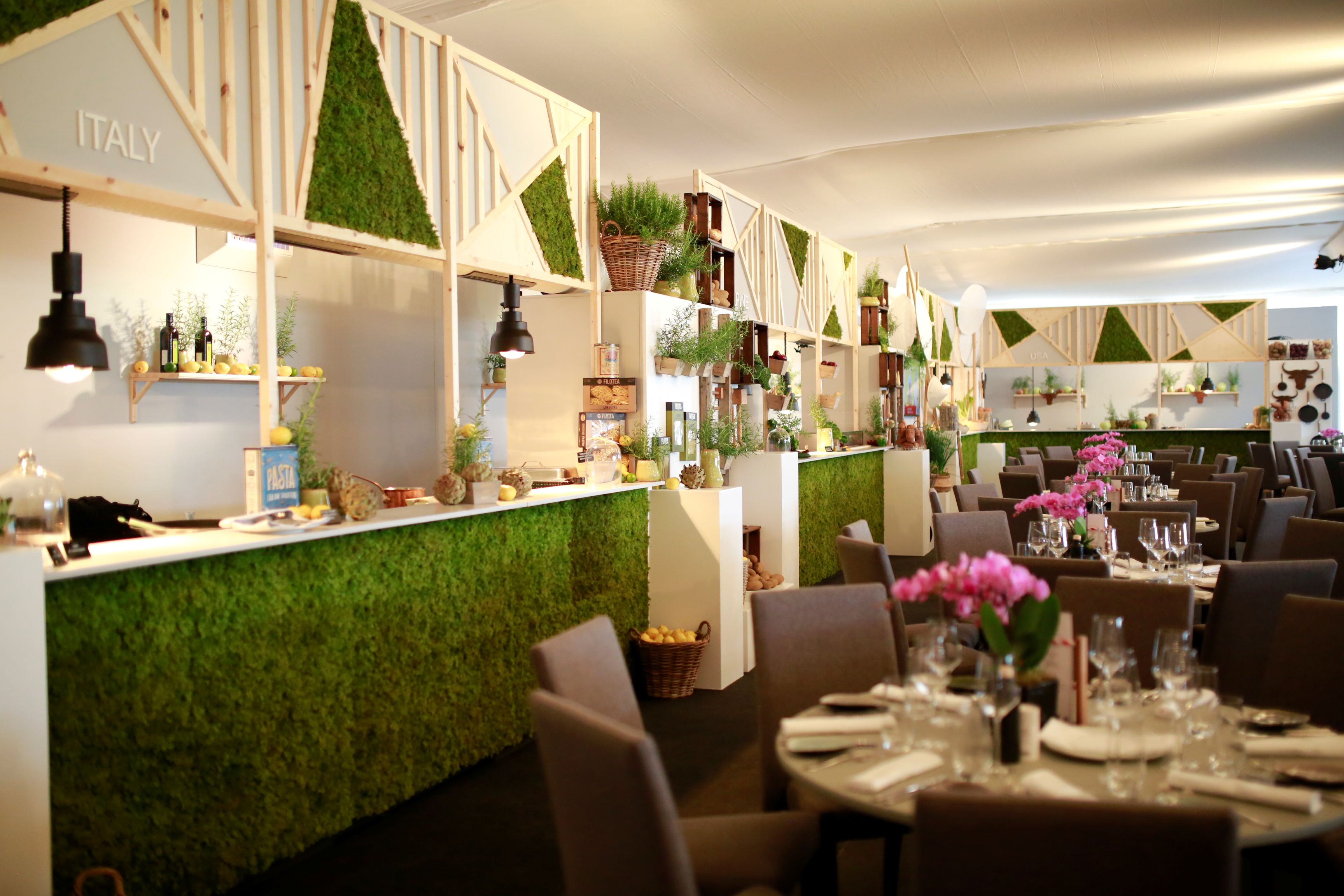 Blick in ein stilvoll dekoriertes Hospitality-Zelt mit langen Reihen gedeckter Tische; entlang der Wand verläuft eine Theke mit grüner Moosverkleidung und Länderschildern wie „Italy“ und „USA“, dazu hängen schwarze Lampen und der Raum ist mit Pflanzen und weiteren Deko-Elementen dekoriert.