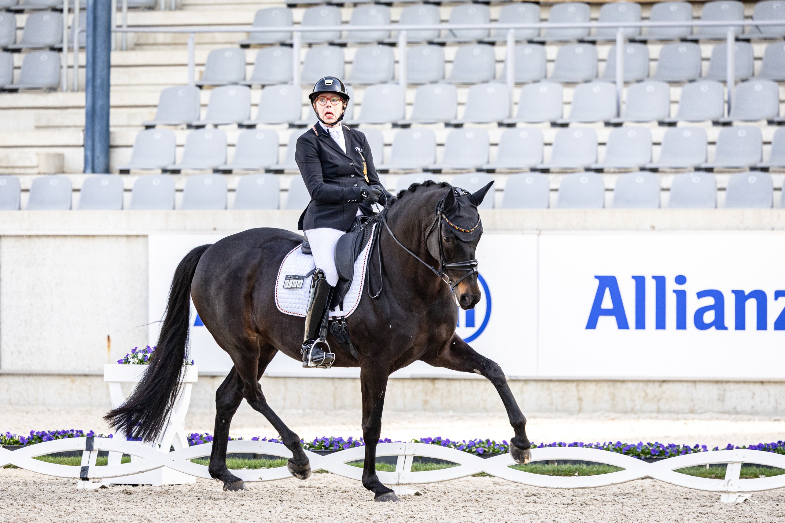 Para dressage rider in a black jacket and helmet on a dark horse performing an extended trot in a sandy arena, with grey grandstand seats and an Allianz advertising banner in the background.