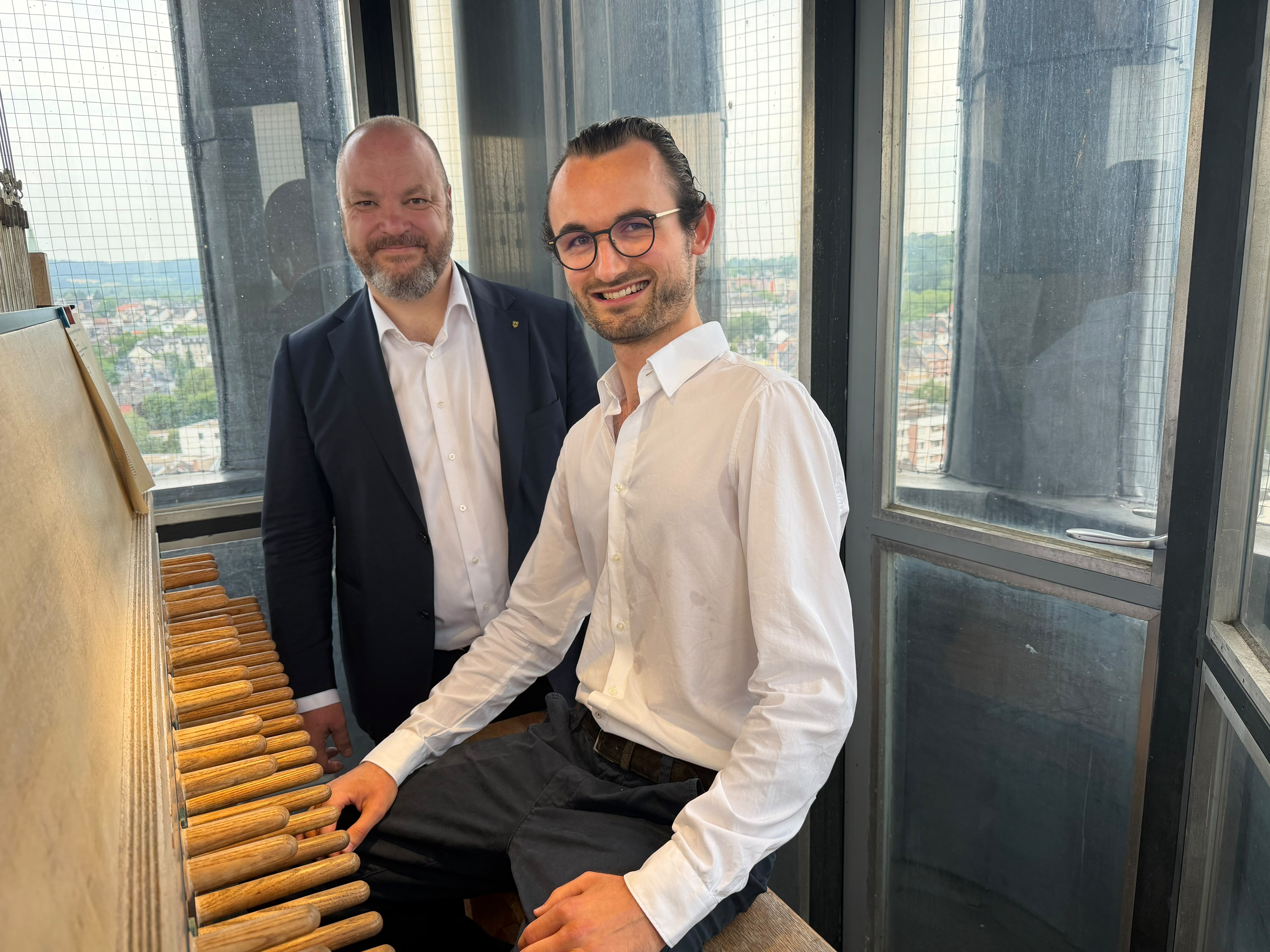 A young man in a white shirt is sitting and smiling at a carillon with many wooden levers, with a man in a dark suit standing next to him; through the surrounding windows, the cityscape of Aachen can be seen in the background.
