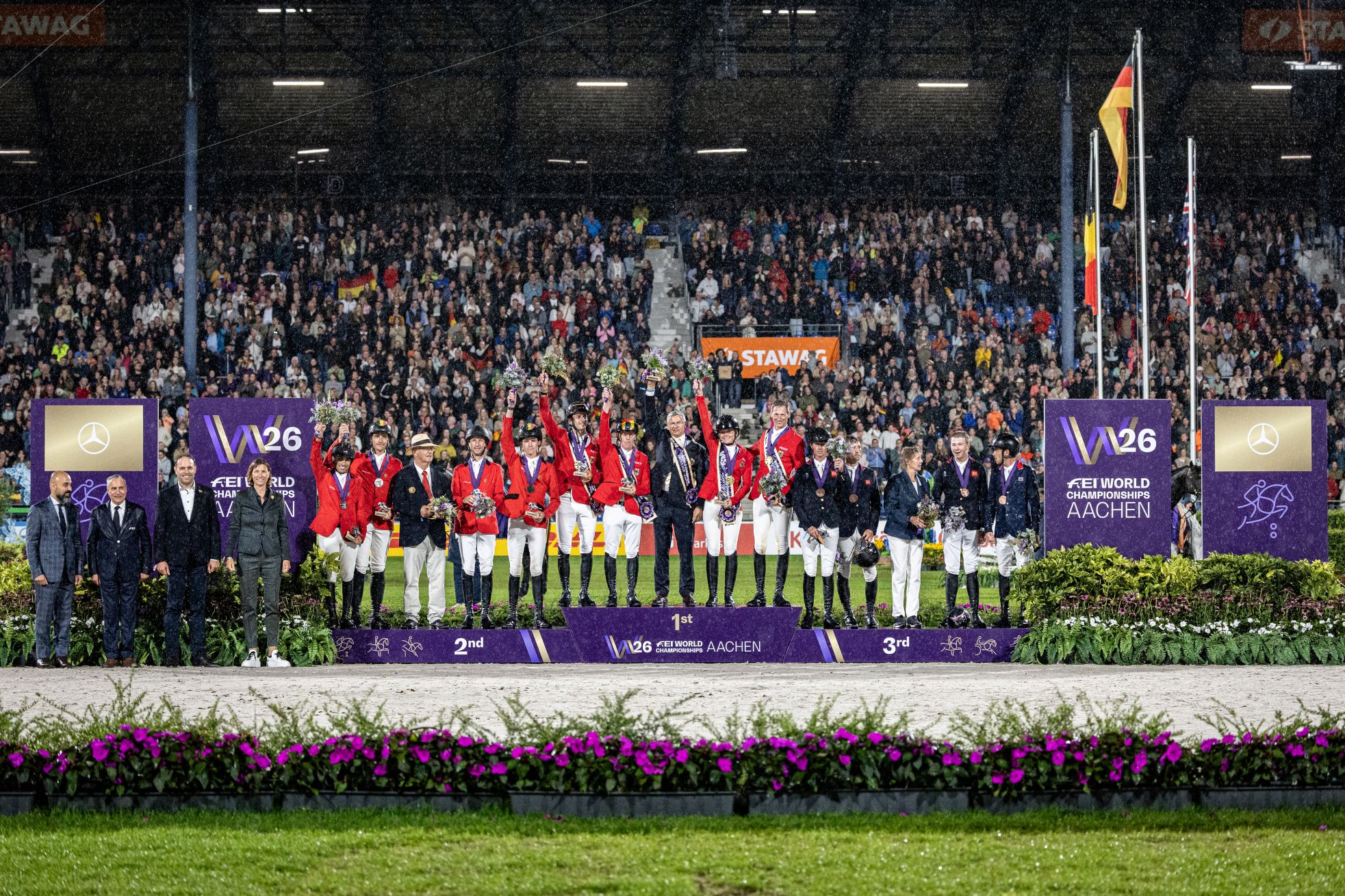 Medal Ceremony at the 2026 FEI World Championships in Aachen in the rain: riders in red and dark blue jackets stand on the podium in first, second and third places, holding medals, flowers and trophies. Event and Mercedes-Benz advertising hoardings line both sides; in the background, a packed grandstand cheers from beneath the arena roof.