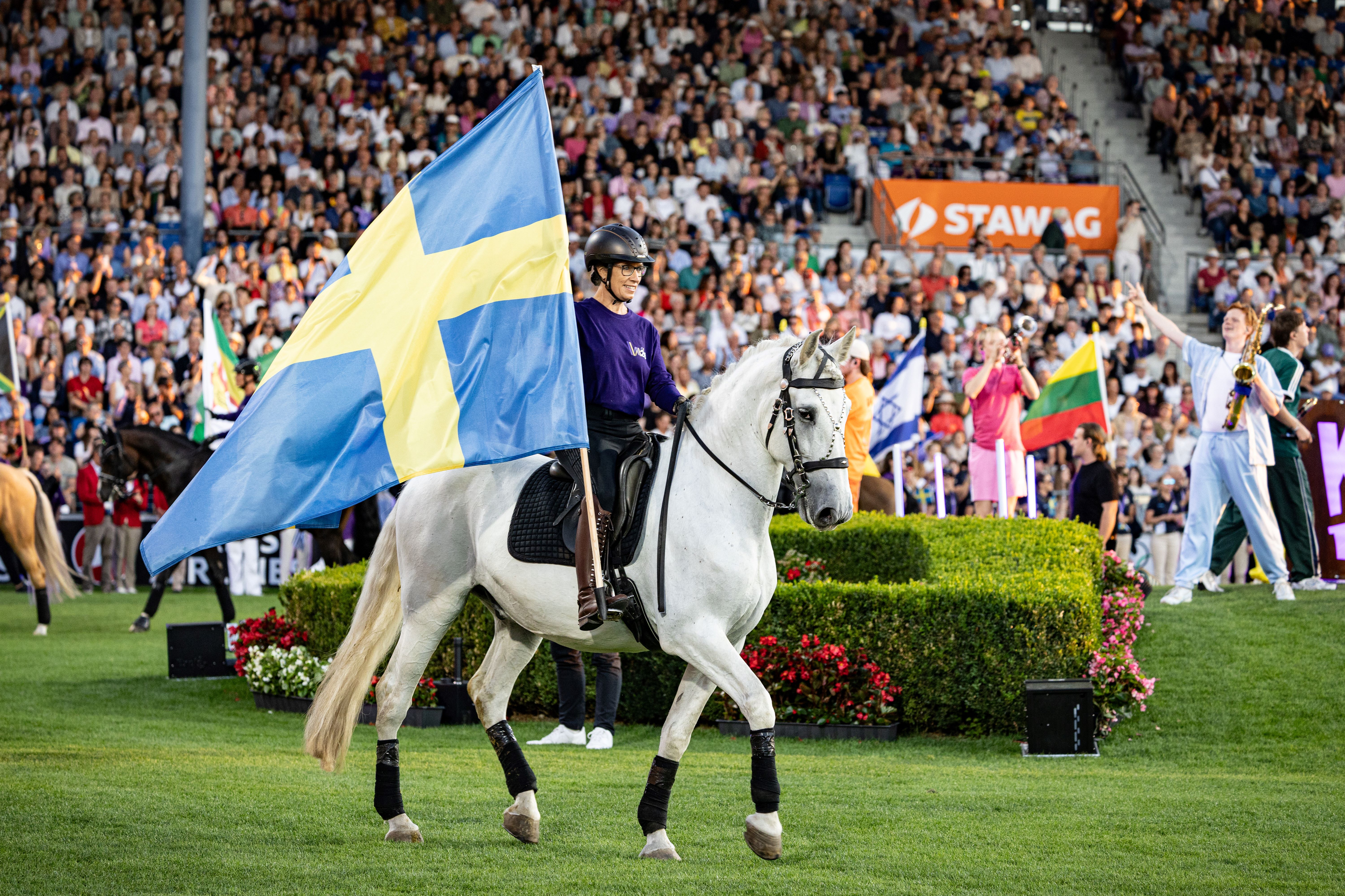 A rider on a white horse carries a large Swedish flag through a packed stadium; in the background are more flags, spectator stands and colourful decorations during the march-in of the nations.