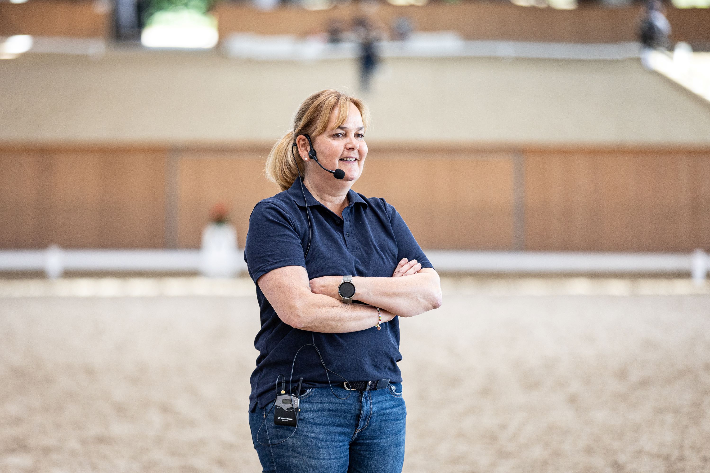A woman wearing a headset microphone and with her arms crossed is standing, smiling, in a brightly lit riding hall against a blurred background.