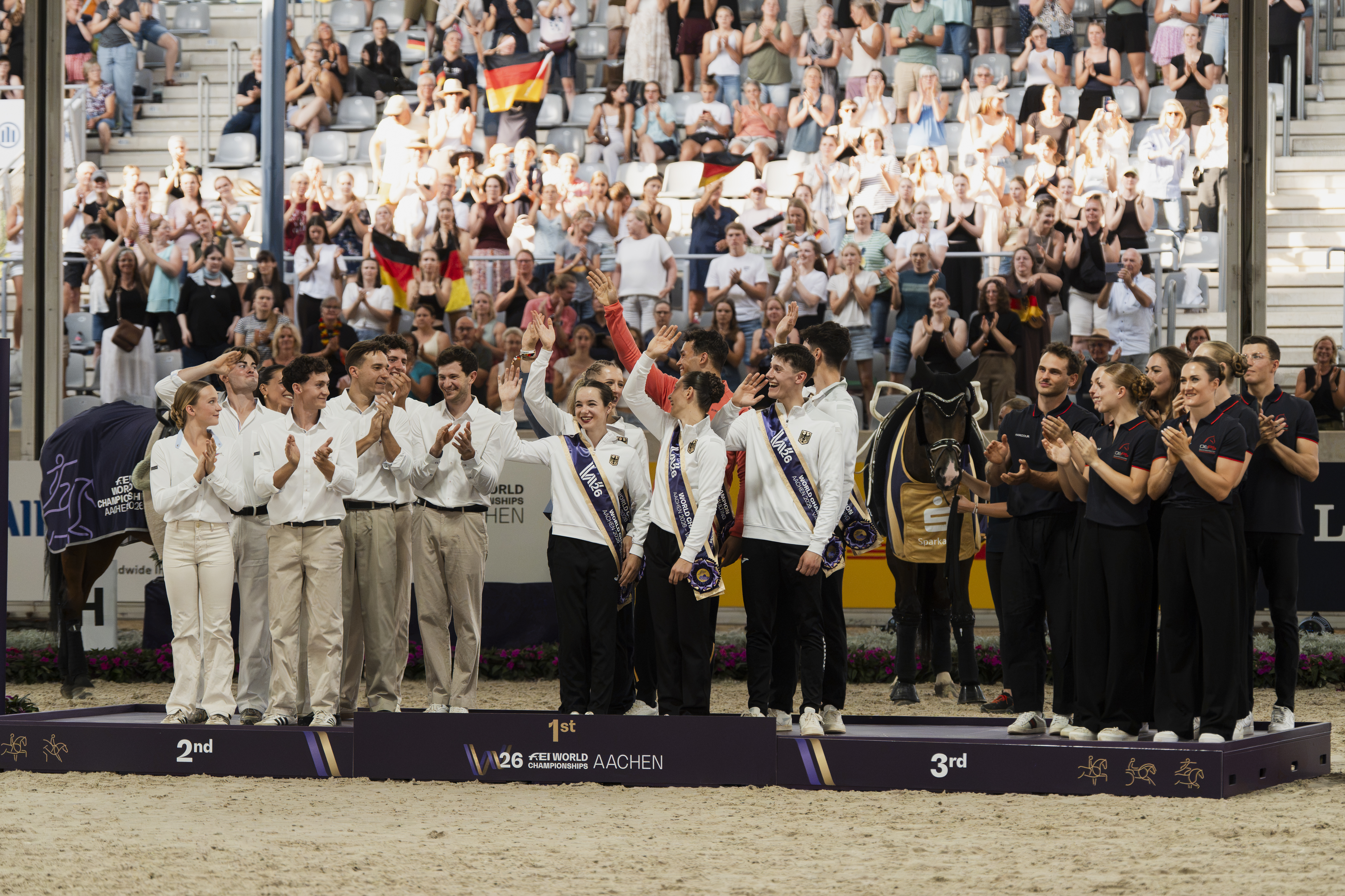 Siegerehrung im Voltigieren: Eine Gruppe junger Voltigierer in weiß-schwarzer Teamkleidung mit Deutschland-Emblem und Schärpen steht jubelnd auf dem Podium, links und rechts von Trainer- und Helferteams flankiert, im Hintergrund ein applaudierendes, mit Deutschland-Fahnen wedelndes Publikum und ein voltigierfertig ausgerüstetes Pferd.