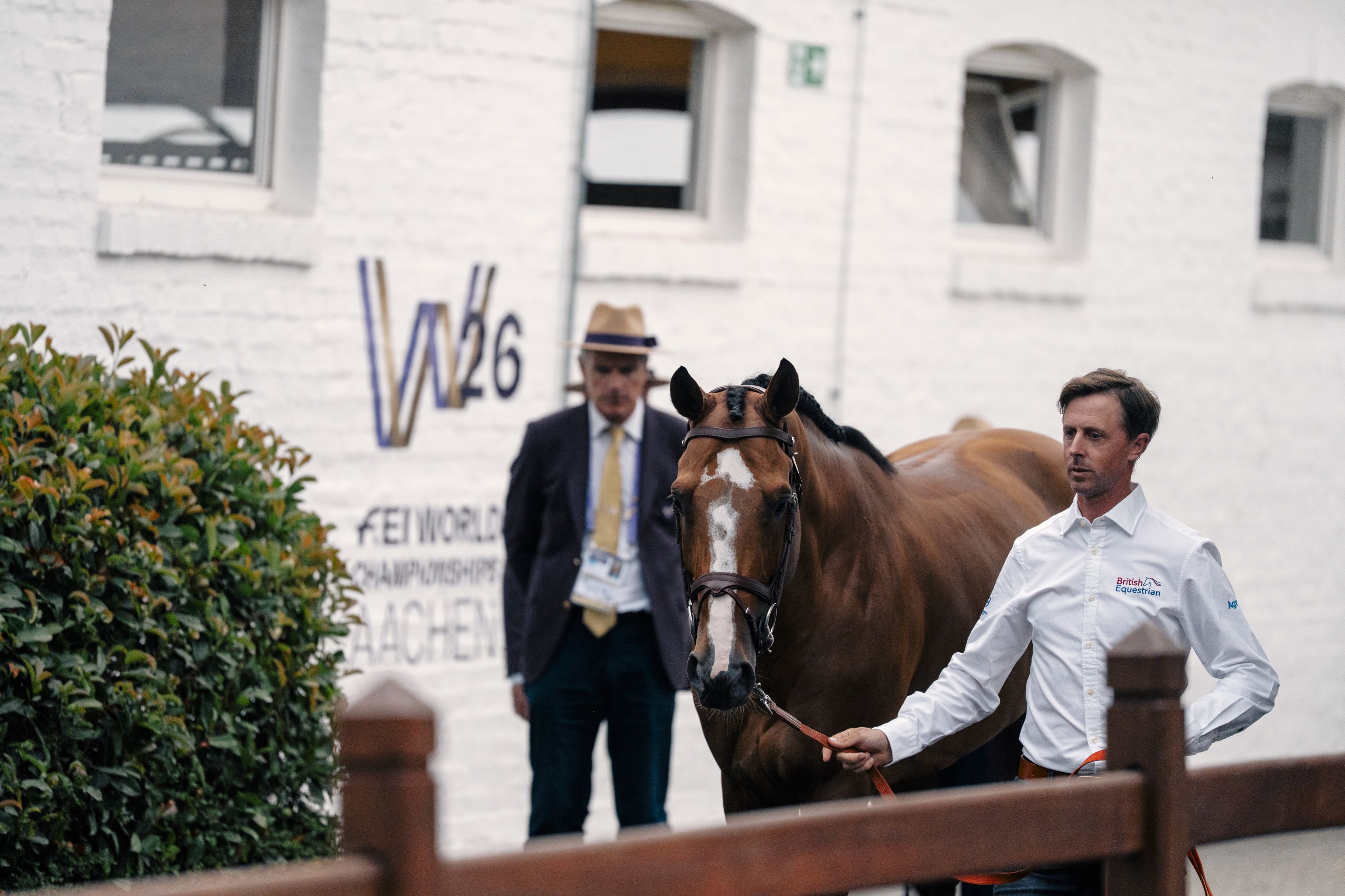 A rider leading a bay horse alongside a wooden fence. A white stable wall displaying the "W26 – FEI World Championships Aachen" logo and an official wearing a suit are visible in the background.
