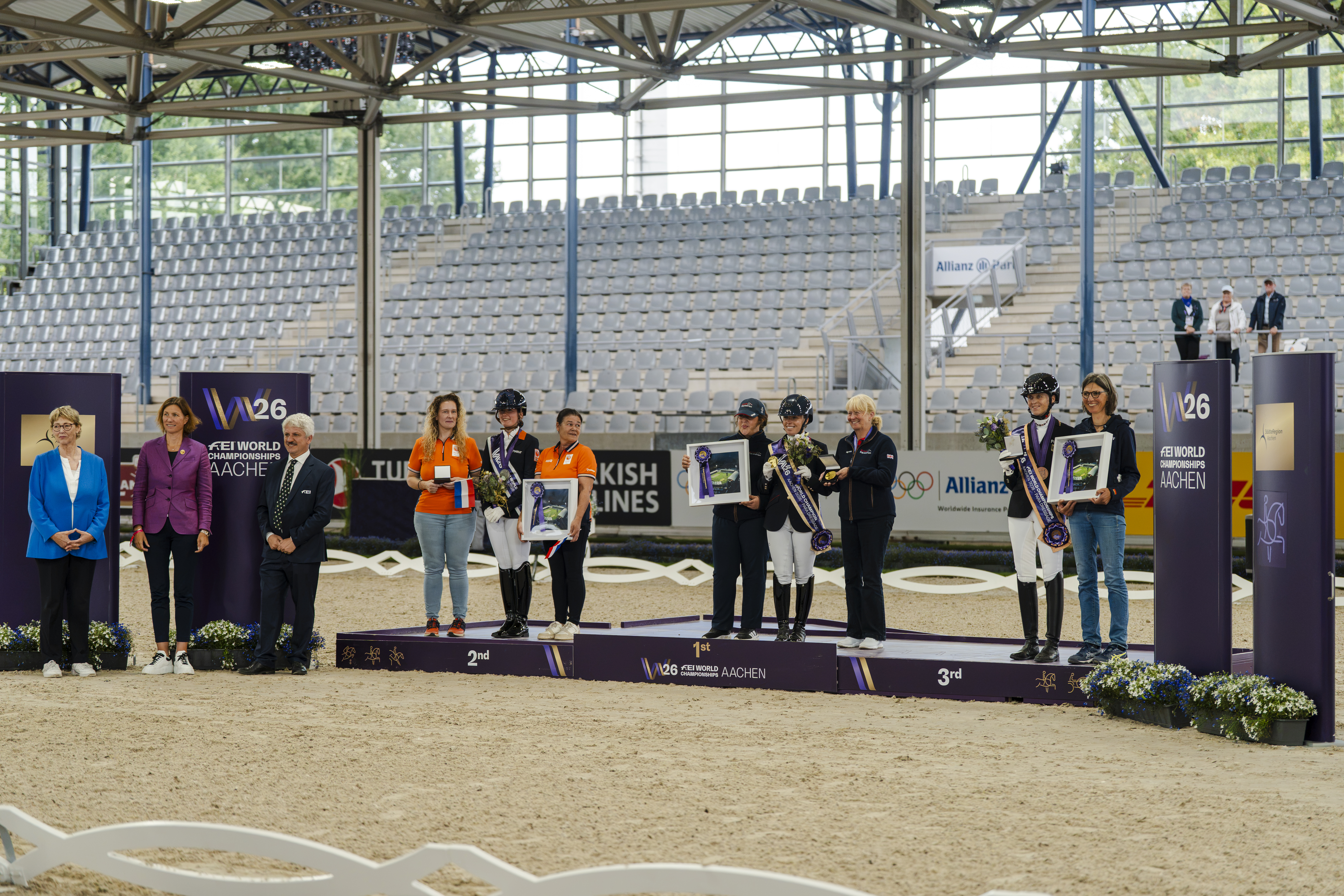Medaillenzeremonie der Para-Dressur bei den FEI-Weltmeisterschaften Aachen 2026: Drei Reiterinnen stehen mit jeweils einer weiteren Person auf den Podesten für den ersten, zweiten und dritten Platz und halten Blumen sowie gerahmte Auszeichnungen. Die Zeremonie findet in einer Reitarena mit Tribünen statt.