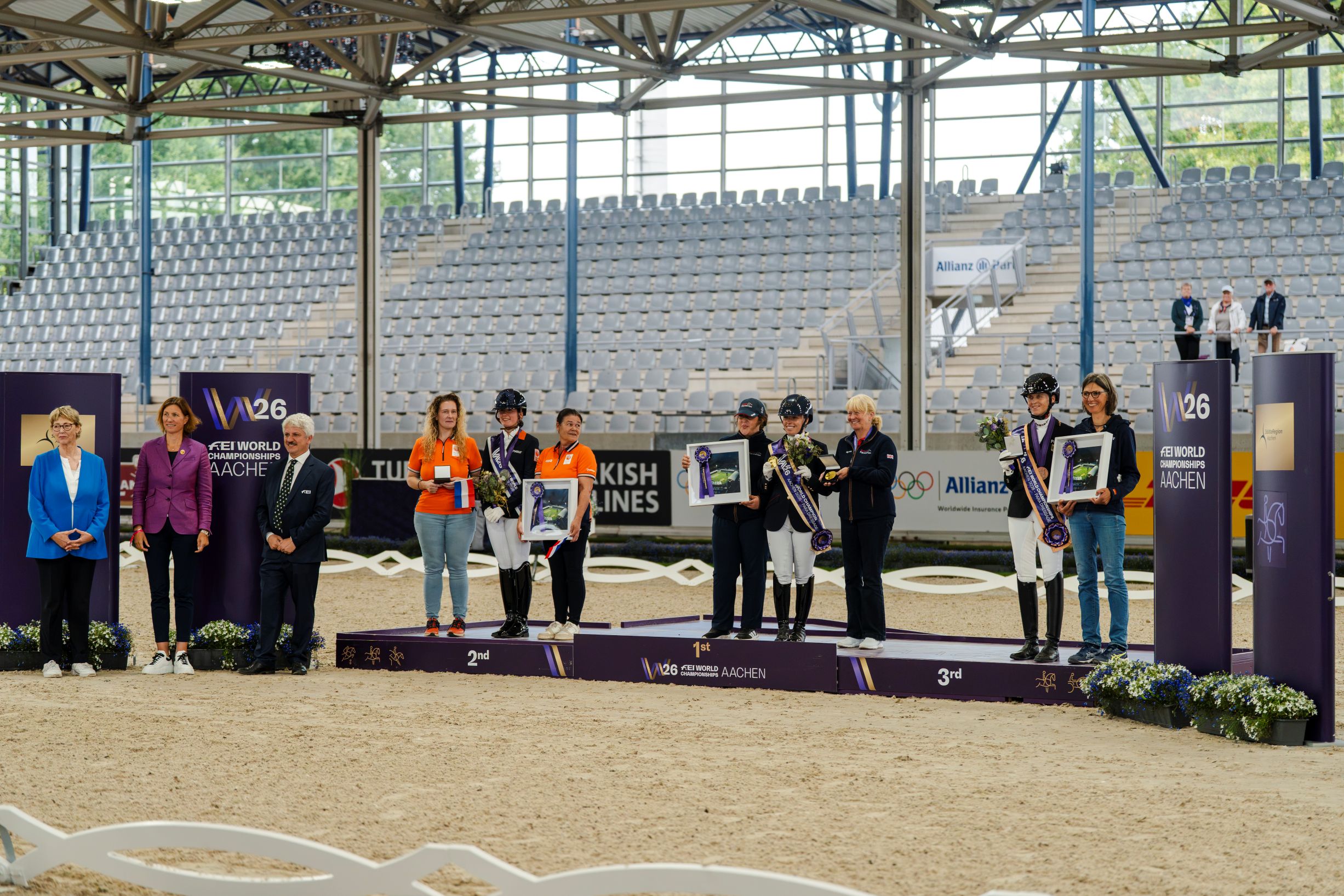 Medal Ceremony for Para Dressage at the 2026 FEI World Championships in Aachen: Three riders, each accompanied by one other person, stand on the podiums for first, second and third place, holding flowers and framed certificates. The ceremony takes place in an equestrian arena with stands.