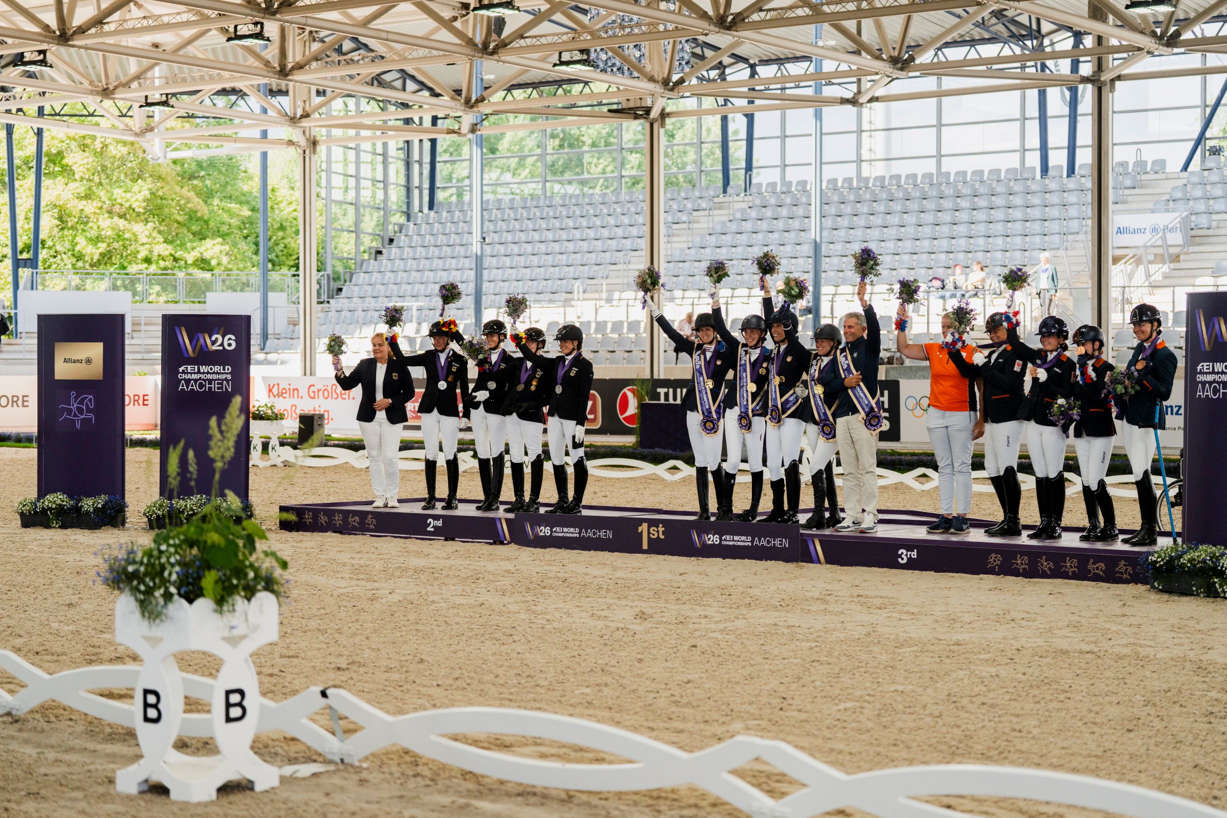 Medal ceremony in Para Dressage: Riders stand on the podium in second, first, and third place, wearing medals, sashes, and holding bouquets of flowers. In the background, you can see the grandstands and signs for the FEI World Championships Aachen 2026.