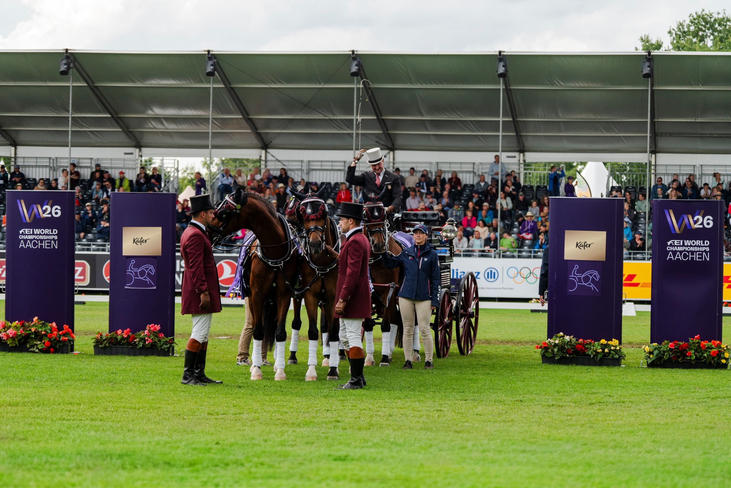 Prize-giving ceremony for the driving event: A coachman in a black suit and white top hat is seated in a carriage behind a team of four bay horses and is raising one hand. Two assistants in burgundy uniforms and another person are standing beside the team. In the background, spectators are watching the ceremony; signs read ‘FEI World Championships Aachen’ and ‘Käfer’.