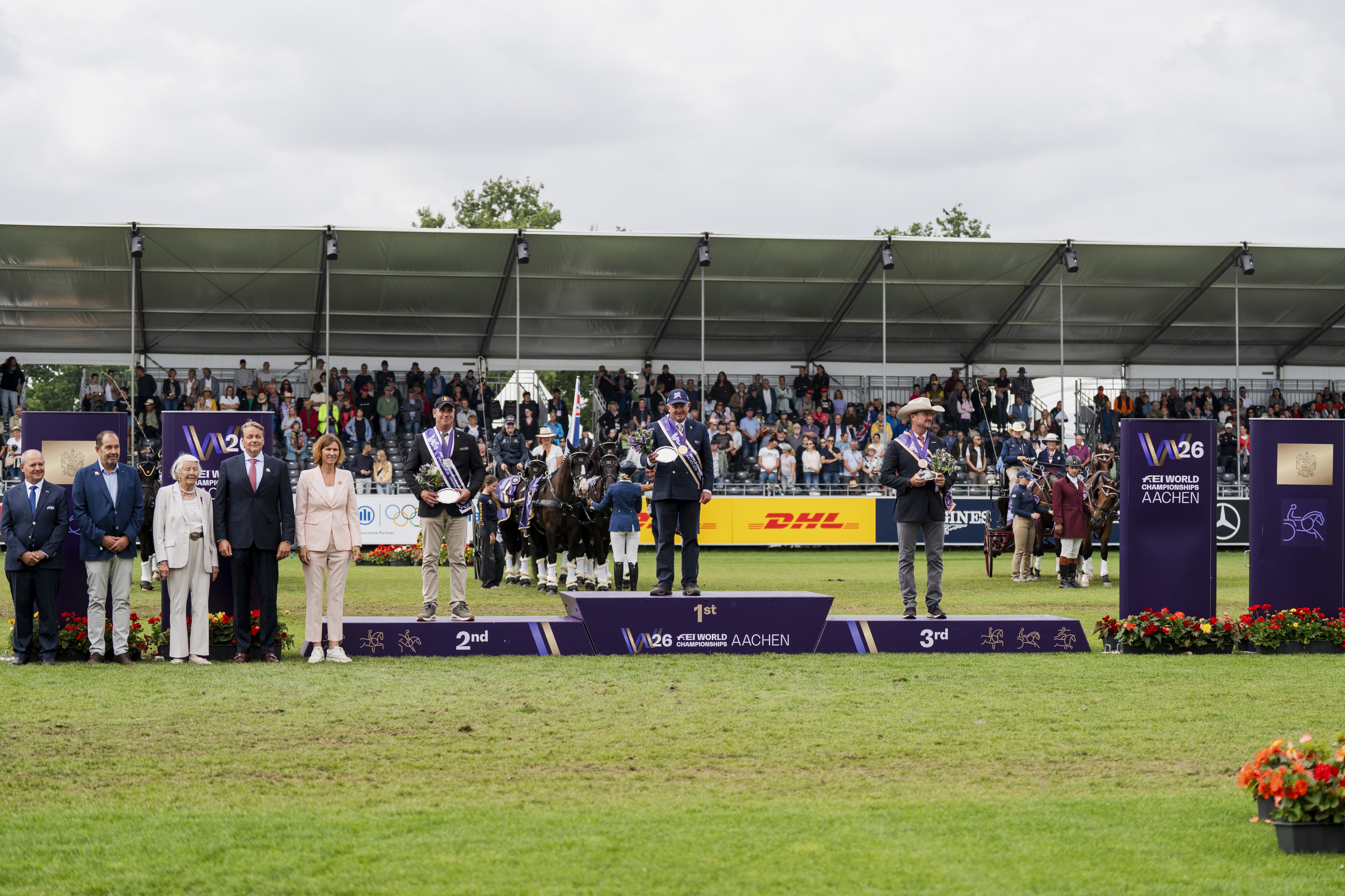 Siegerehrung im Gespannfahren bei den FEI World Championships Aachen 2026: Drei ausgezeichnete Fahrer stehen mit ihren Pferdegespannen auf dem Podium, umgeben von Offiziellen und vor voll besetzten Zuschauertribünen.
