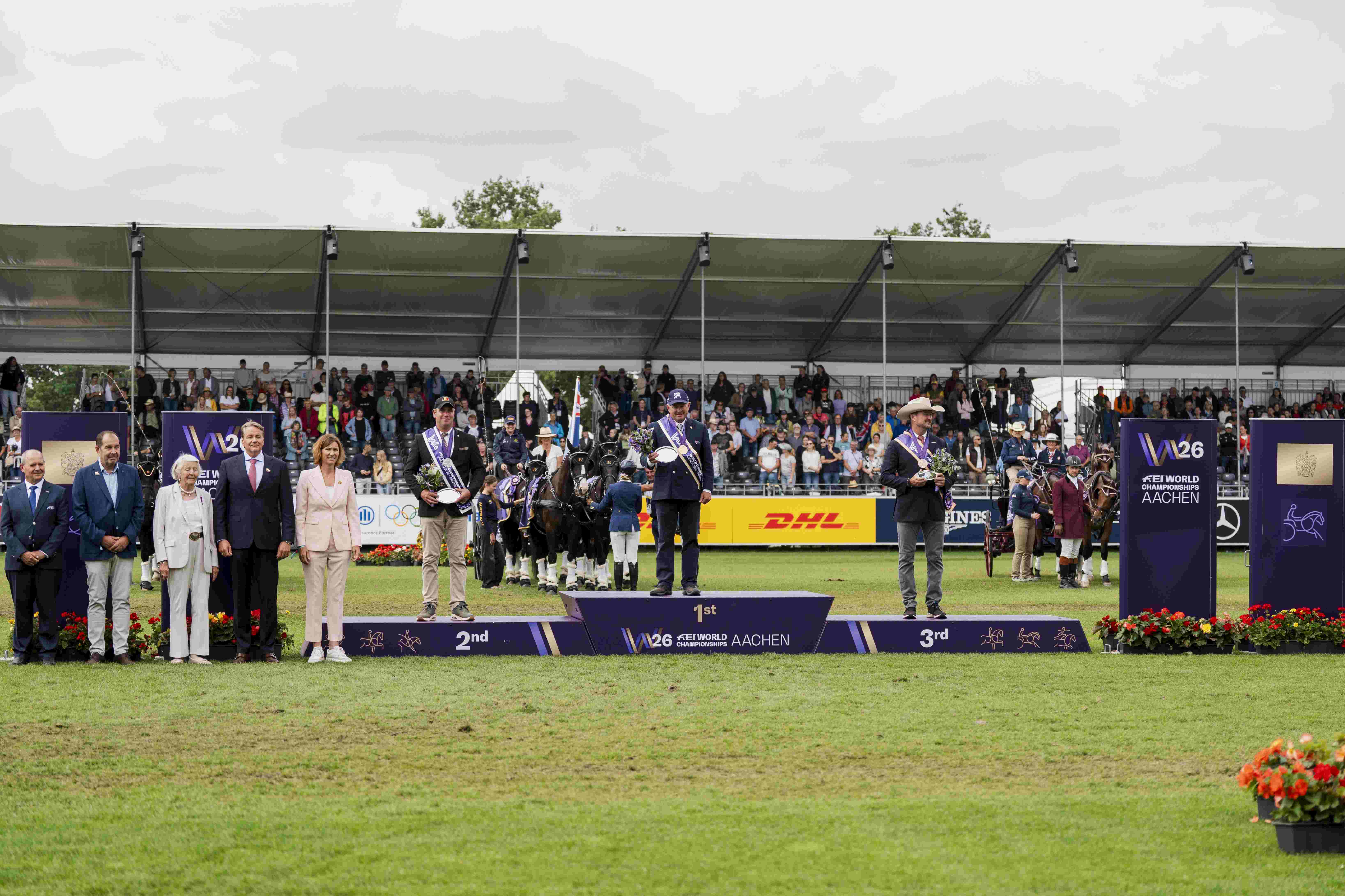 The award ceremony for the FEI Driving World Championship Aachen 2026: three drivers stand on the podium with their horse-drawn carriages, surrounded by officials and in front of packed stands.