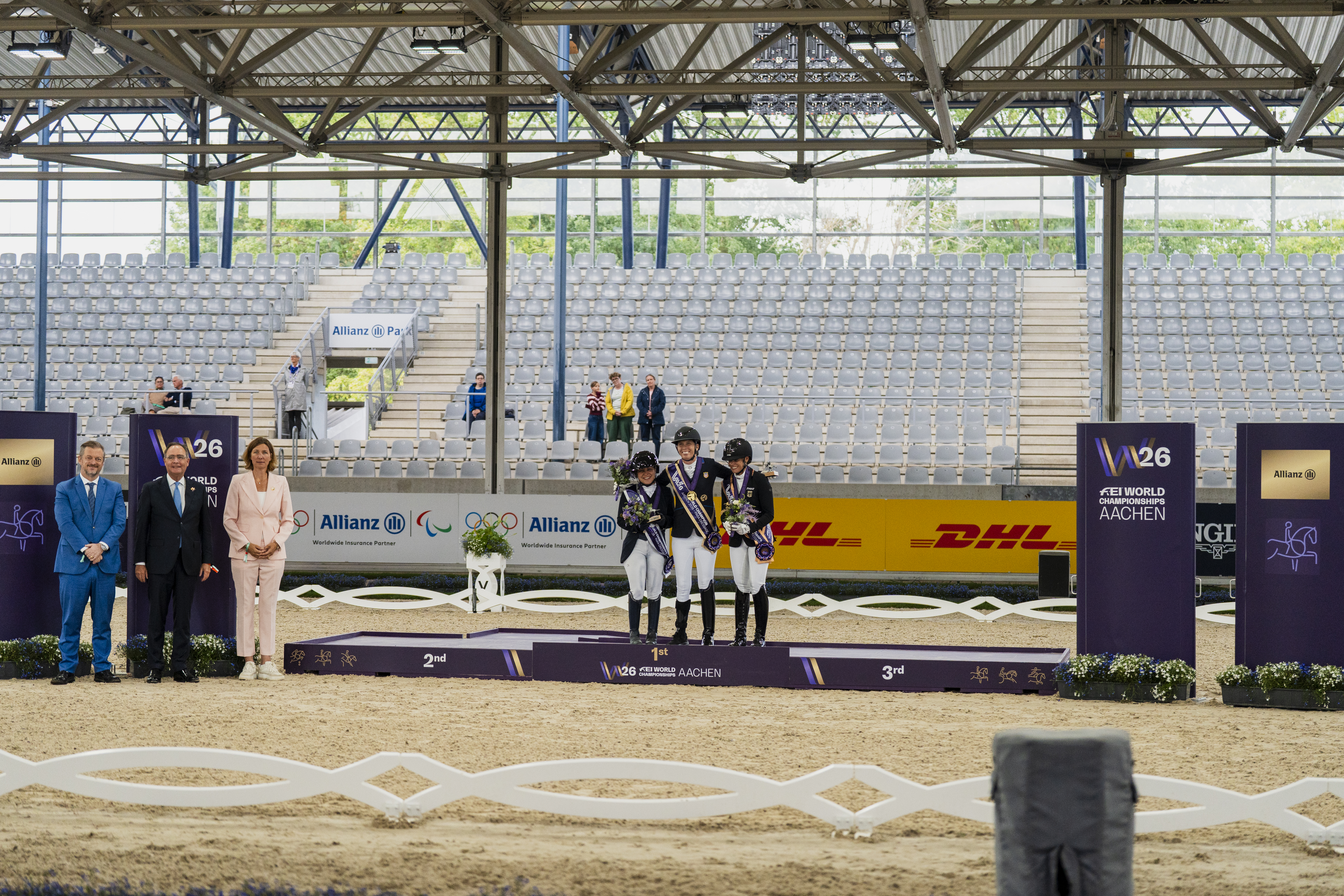 Siegerehrung in der Para-Dressur in Grade IV bei den FEI World Championships Aachen 2026: Drei ausgezeichnete Reiterinnen und Reiter stehen mit Medaillen und Blumen auf dem Podium, begleitet von Ehrengästen in einem Reitstadion.