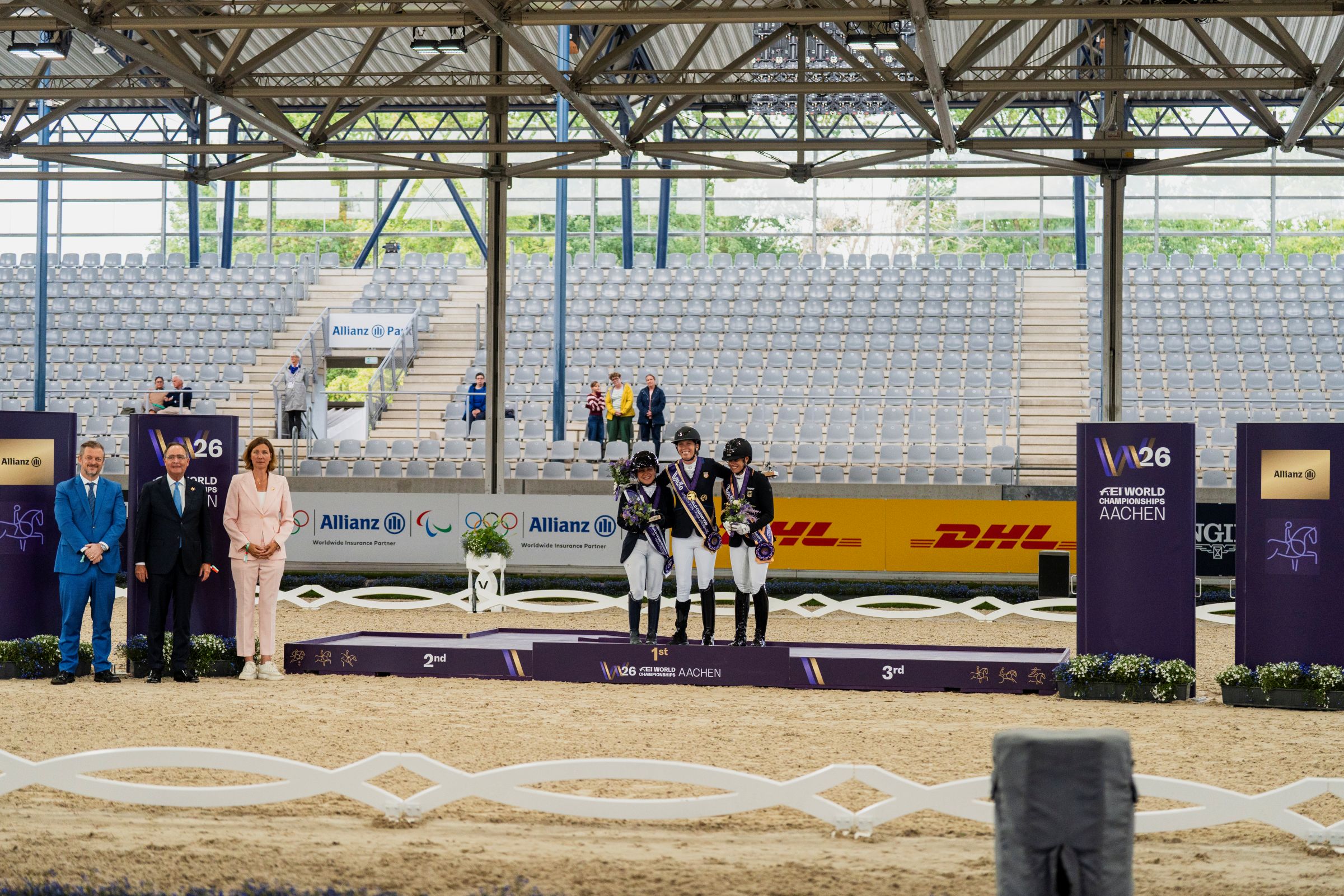 Medal Ceremony for Grade IV Para Dressage at the 2026 FEI World Championships in Aachen: Three outstanding riders stand on the podium with their medals and bouquets, accompanied by guests of honour in an equestrian stadium.