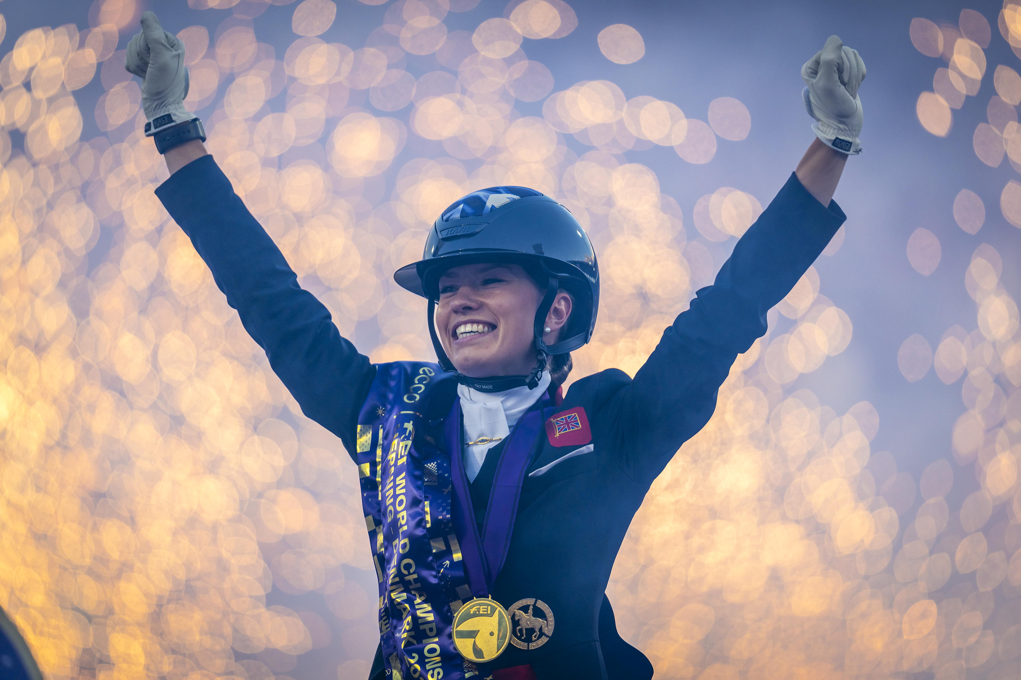 A beaming rider wearing a helmet raises both arms in celebration; she is wearing a blue ‘FEI World Championships’ sash and several gold medals, whilst in the background, lights that appear blurred are glowing.