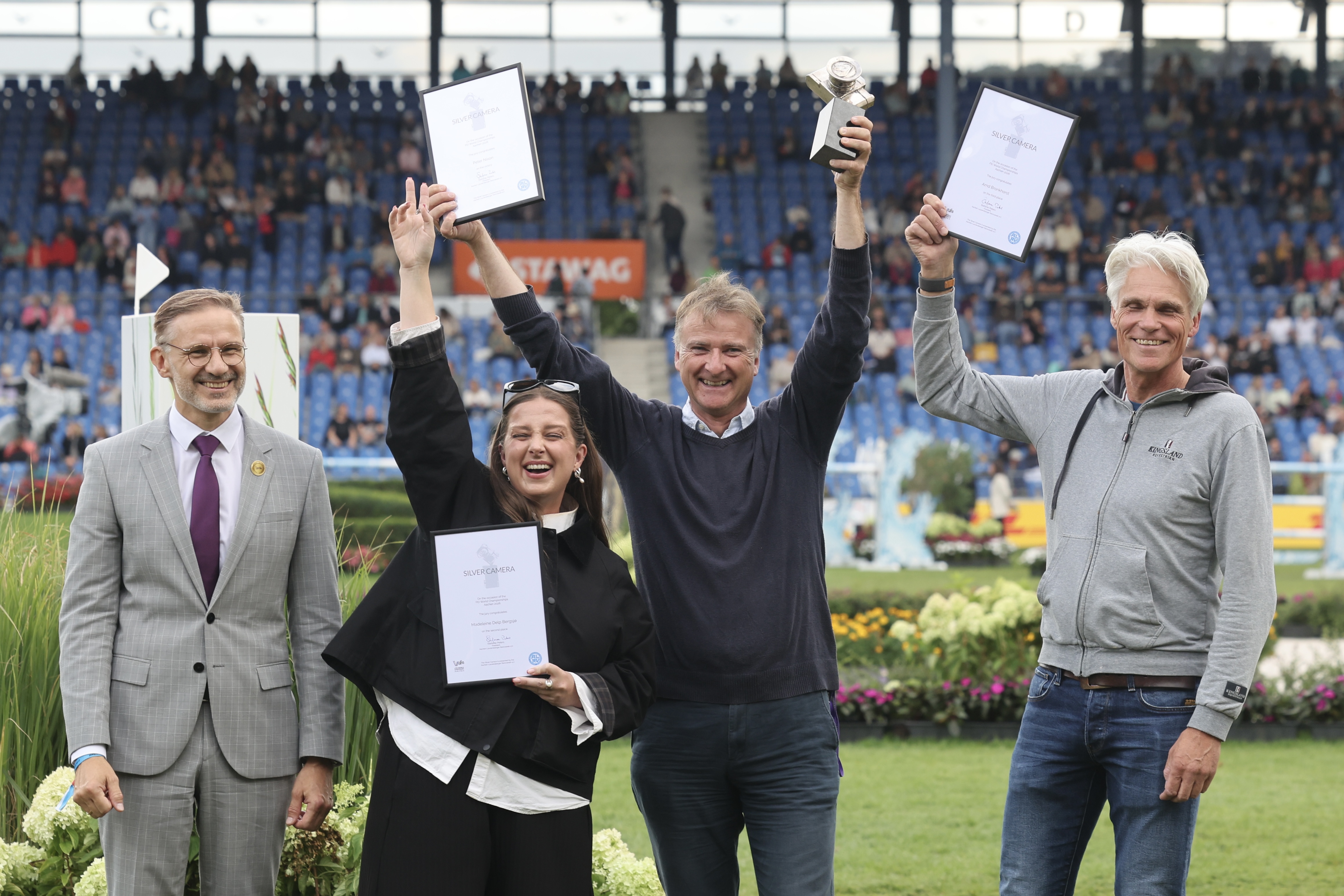 Vier Personen präsentieren bei einer Preisverleihung im Reitstadion ihre Auszeichnungen. Eine Frau und zwei Männer halten gerahmte Urkunden mit der Aufschrift „Silver Camera“ hoch, während ein weiterer Mann eine Trophäe präsentiert. Im Hintergrund sitzen Zuschauer auf den Tribünen.