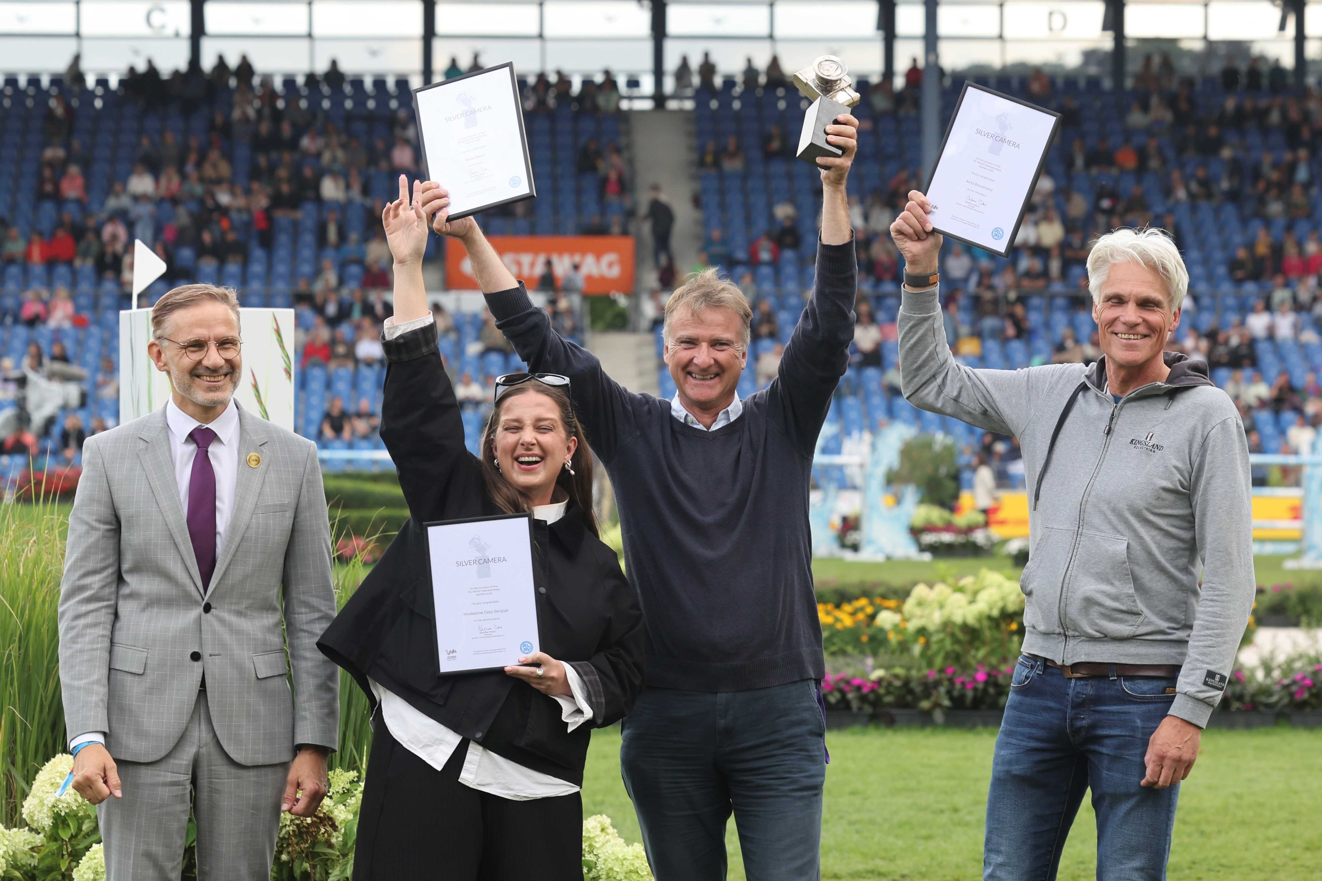 Four people are presenting their awards at a ceremony in the Main Stadium. A woman and two men are holding up framed certificates bearing the words ‘Silver Camera’, whilst another man is presenting a trophy. In the background, spectators are seated in the stands.